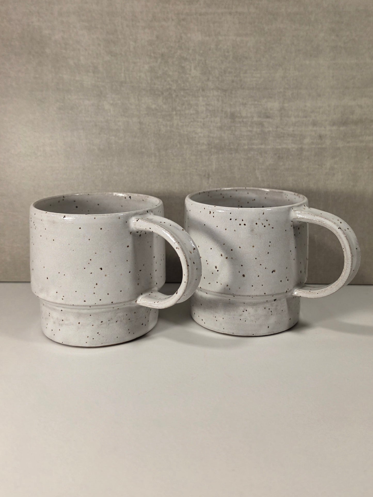 Two handmade ceramic mugs with a speckled light gray and white glaze, displayed side-by-side on a light gray surface against a textured gray background.