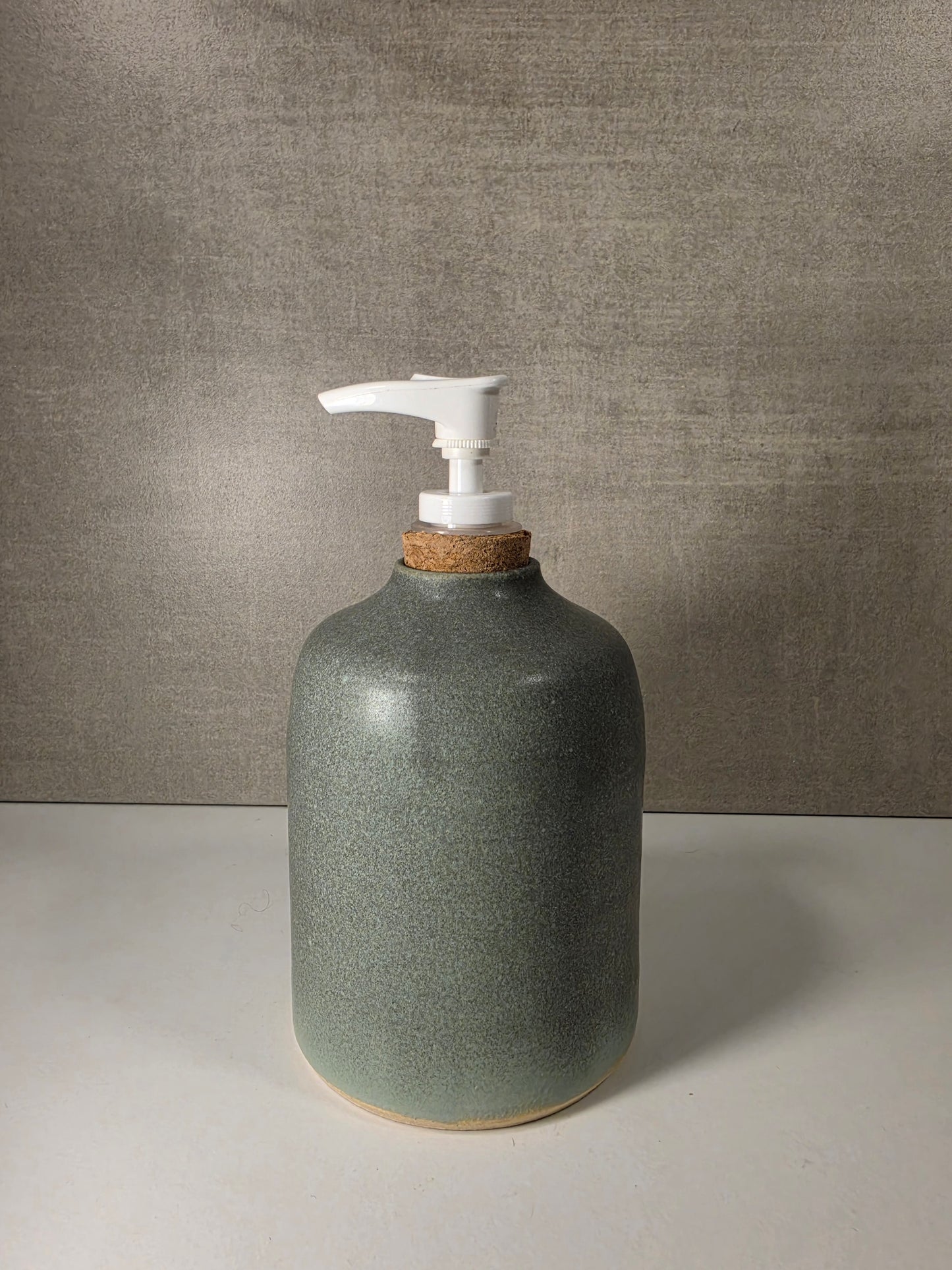 A ceramic soap dispenser bottle with a speckled sage green glaze, a white pump dispenser, and a natural cork stopper, set against a textured gray background.