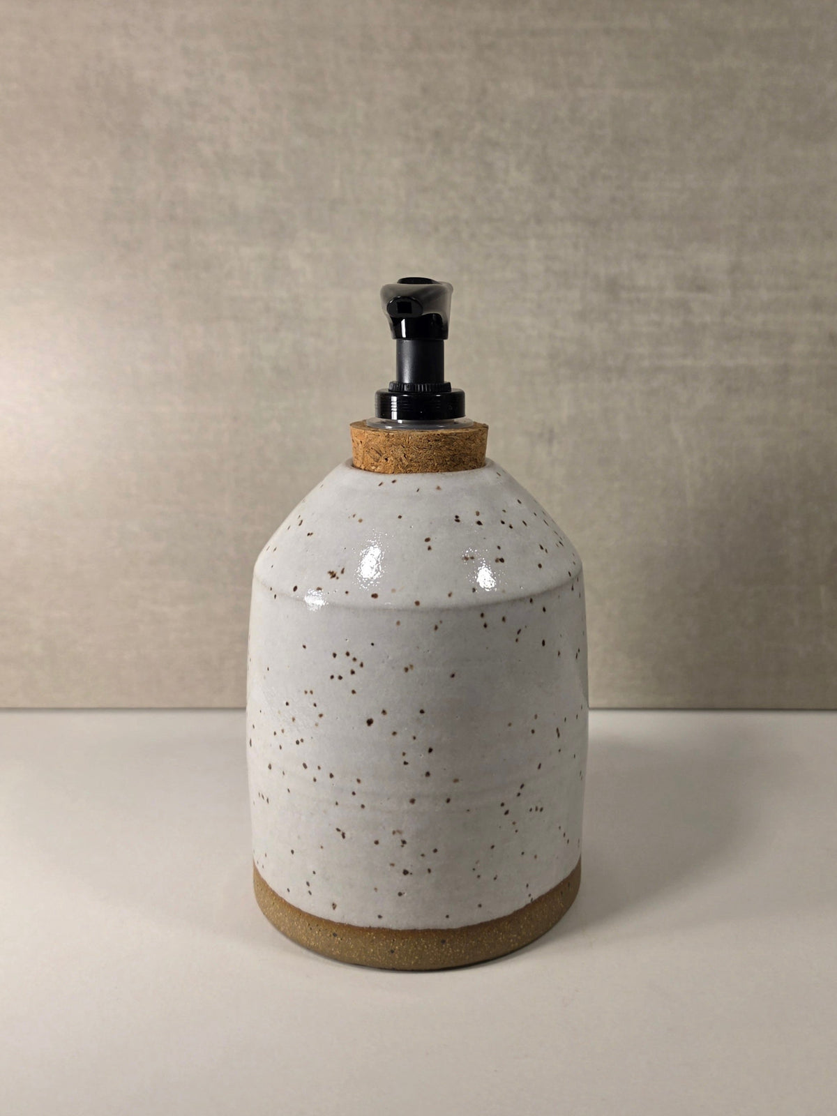 A handmade ceramic soap dispenser bottle with a speckled white glaze, a natural cork stopper, and a black plastic pump, set against a neutral textured beige background.