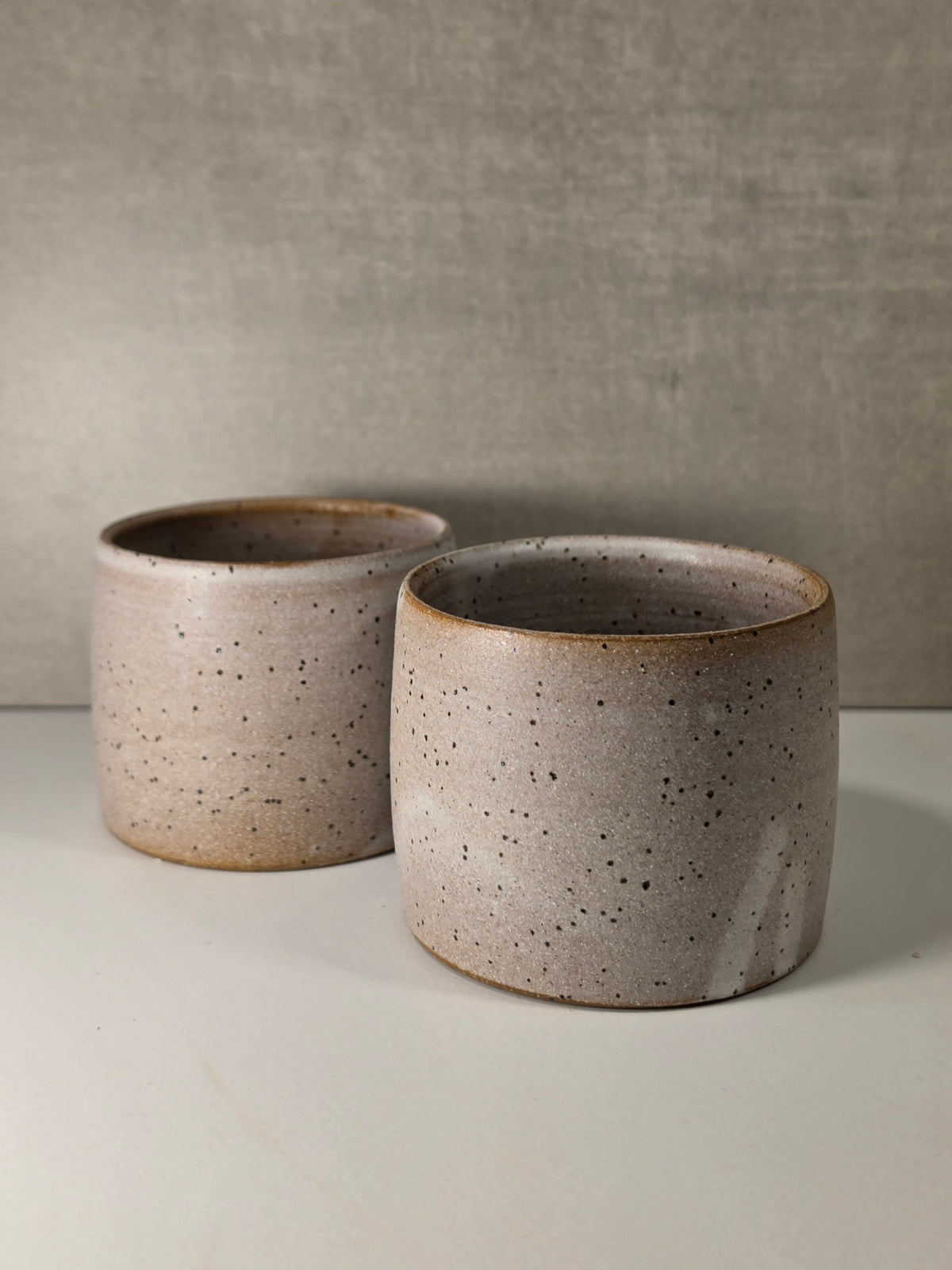 Two small round ceramic cups / mugs with a speckled beige and gray glaze, displayed on a light surface against a textured gray background.