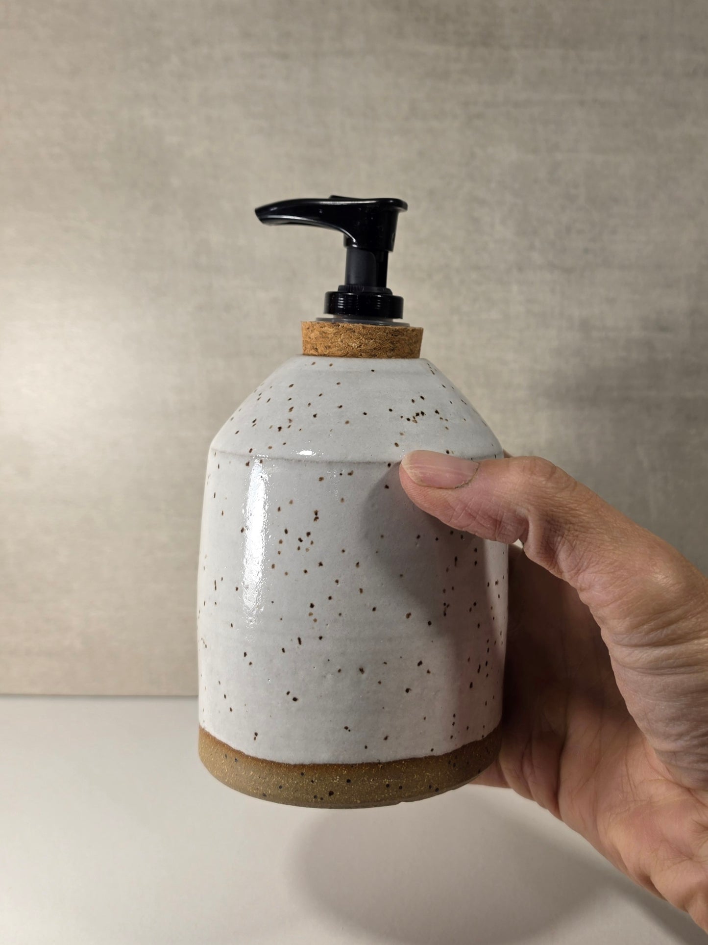 A hand holding a handmade ceramic soap dispenser with a speckled off-white glaze, cork accents, and a black pump, set against a neutral textured beige background.