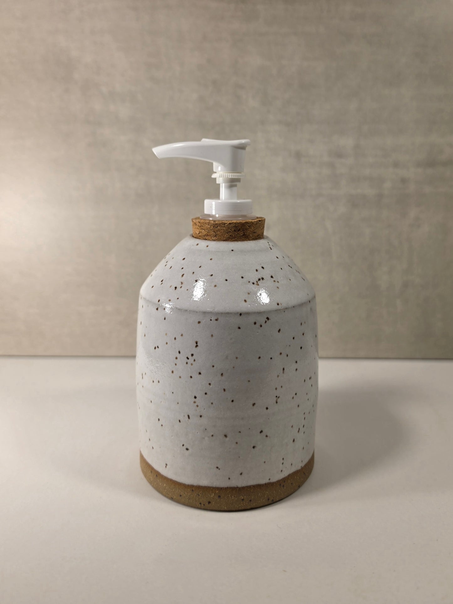 A handmade ceramic soap dispenser with a speckled off-white glaze, a cork neck ring, and a white plastic pump, set against a neutral textured beige background.