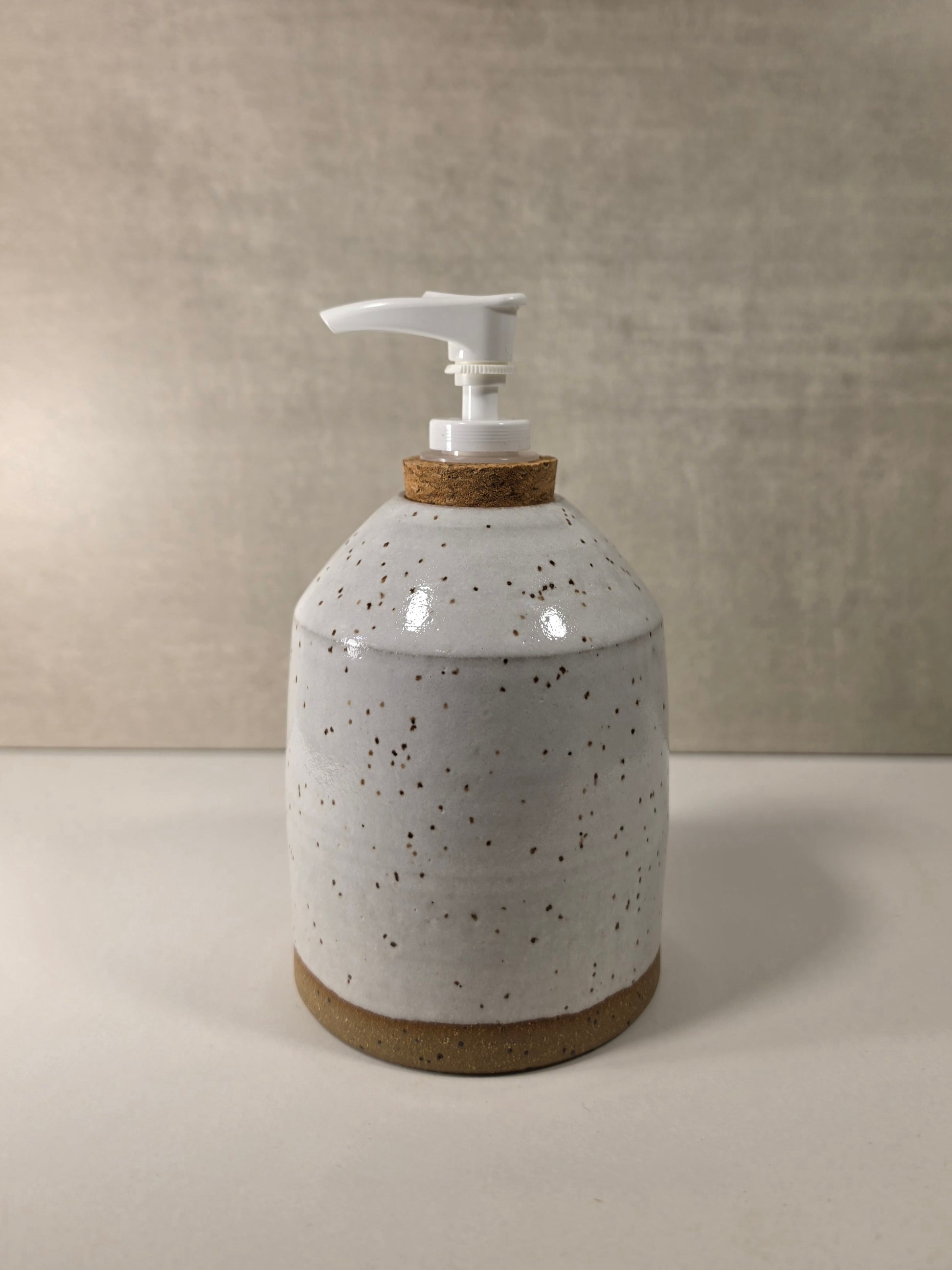 A handmade ceramic soap dispenser with a speckled off-white glaze, a cork neck ring, and a white plastic pump, set against a neutral textured beige background.