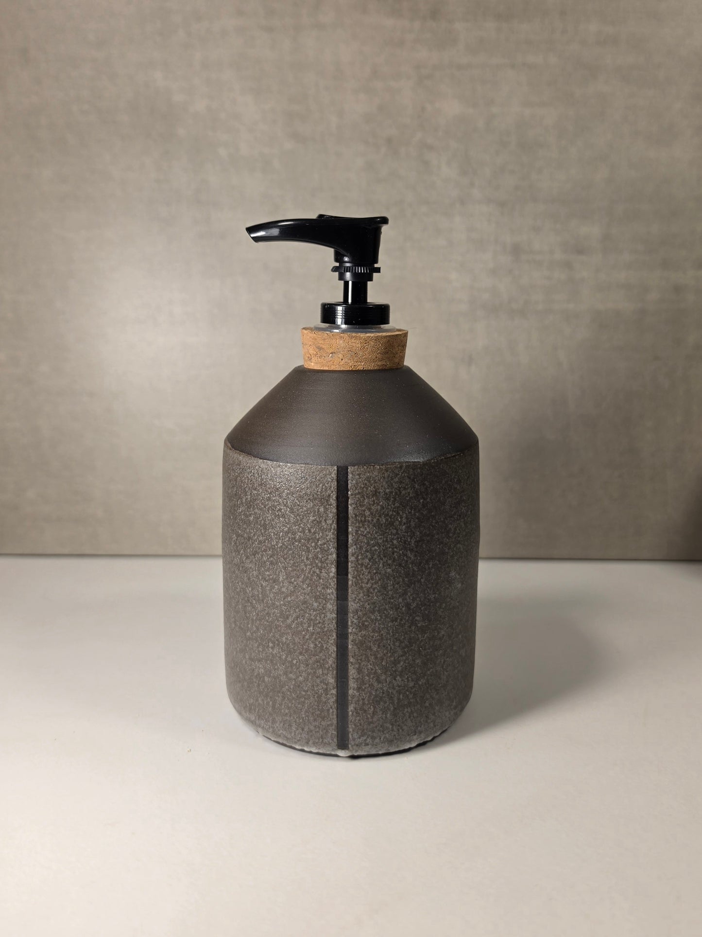 A dark gray textured ceramic soap dispenser with a black pump and a natural cork neck, set against a neutral gray background.