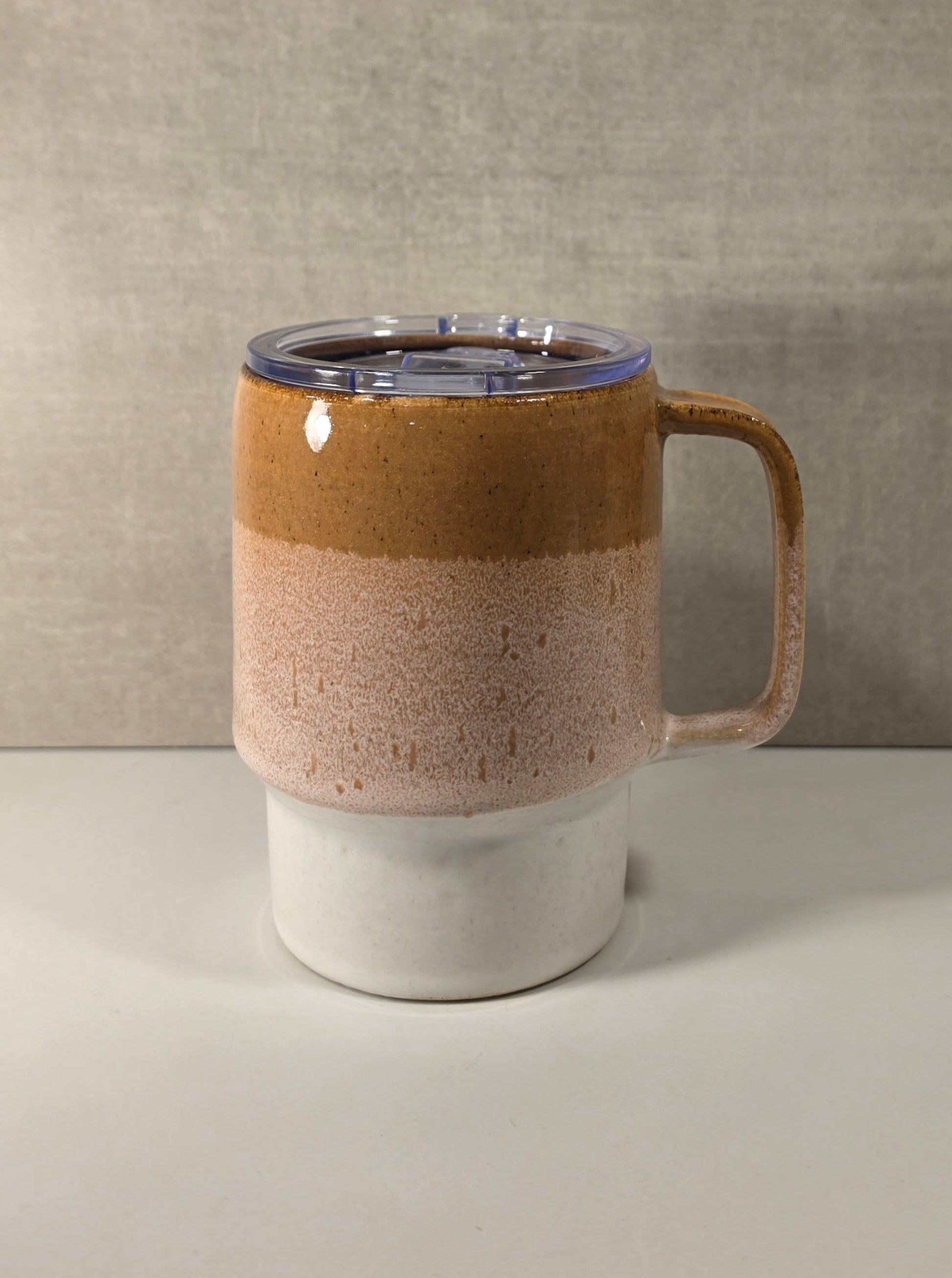 A ceramic travel mug with a handle, featuring a three-layered glaze design in brown, speckled beige, and white, topped with a clear lid.
