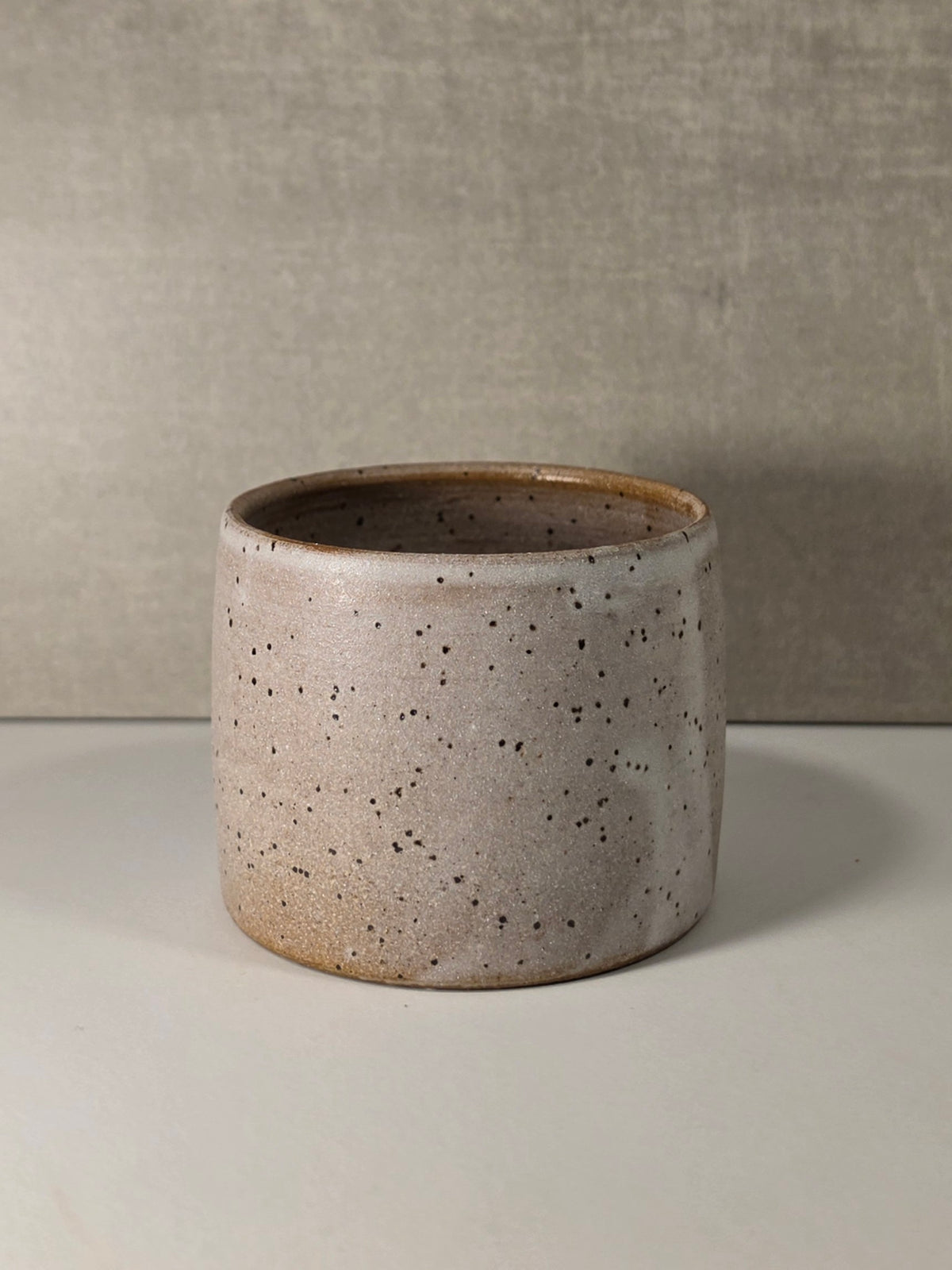 A handmade ceramic artisan cup with a speckled beige and brown glaze, set against a neutral textured gray background.