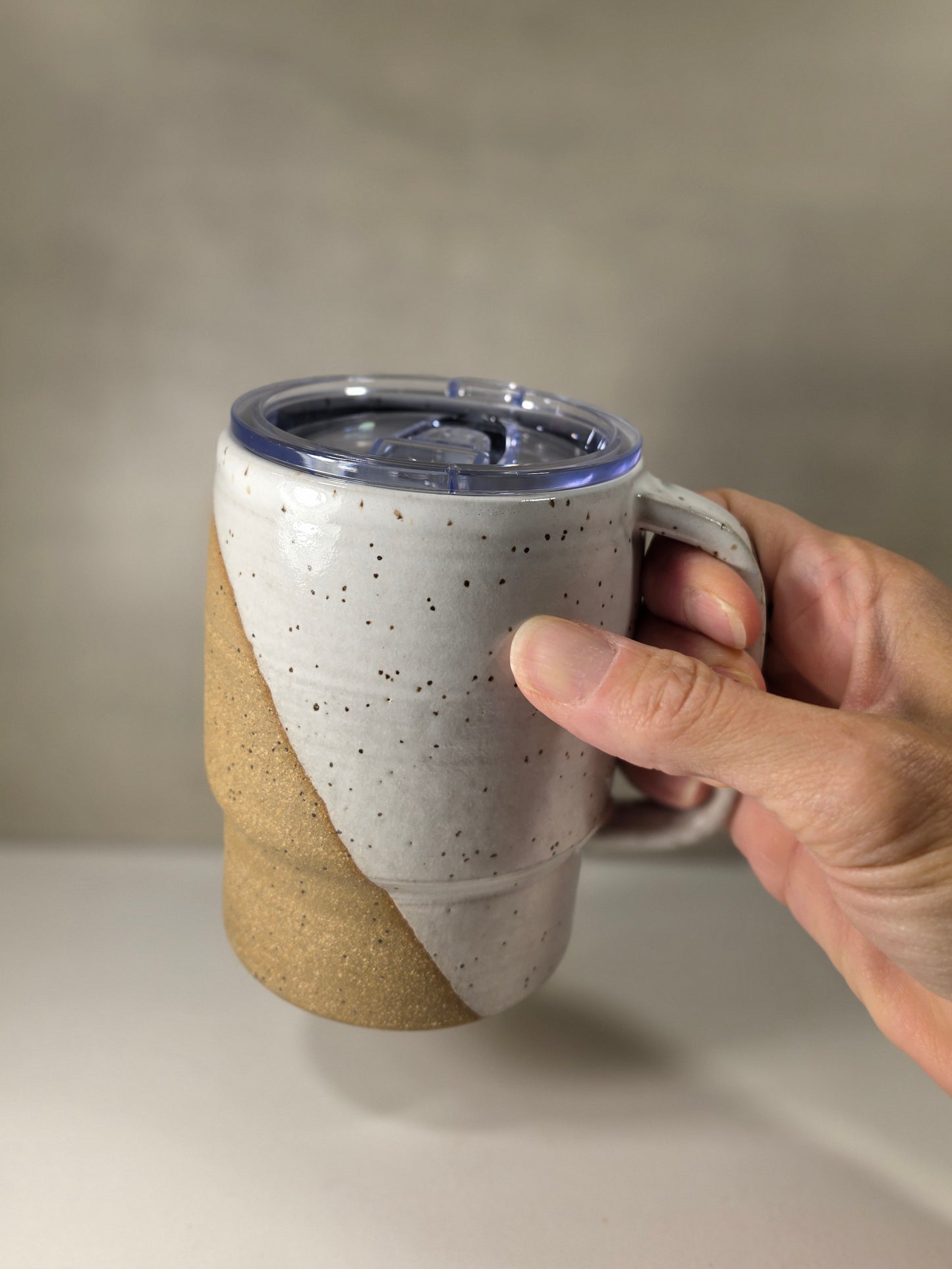 A hand holding a ceramic travel mug with a clear plastic lid. The mug features a dual-tone design with a speckled off-white upper half and a textured tan lower half, set against a neutral gray background.