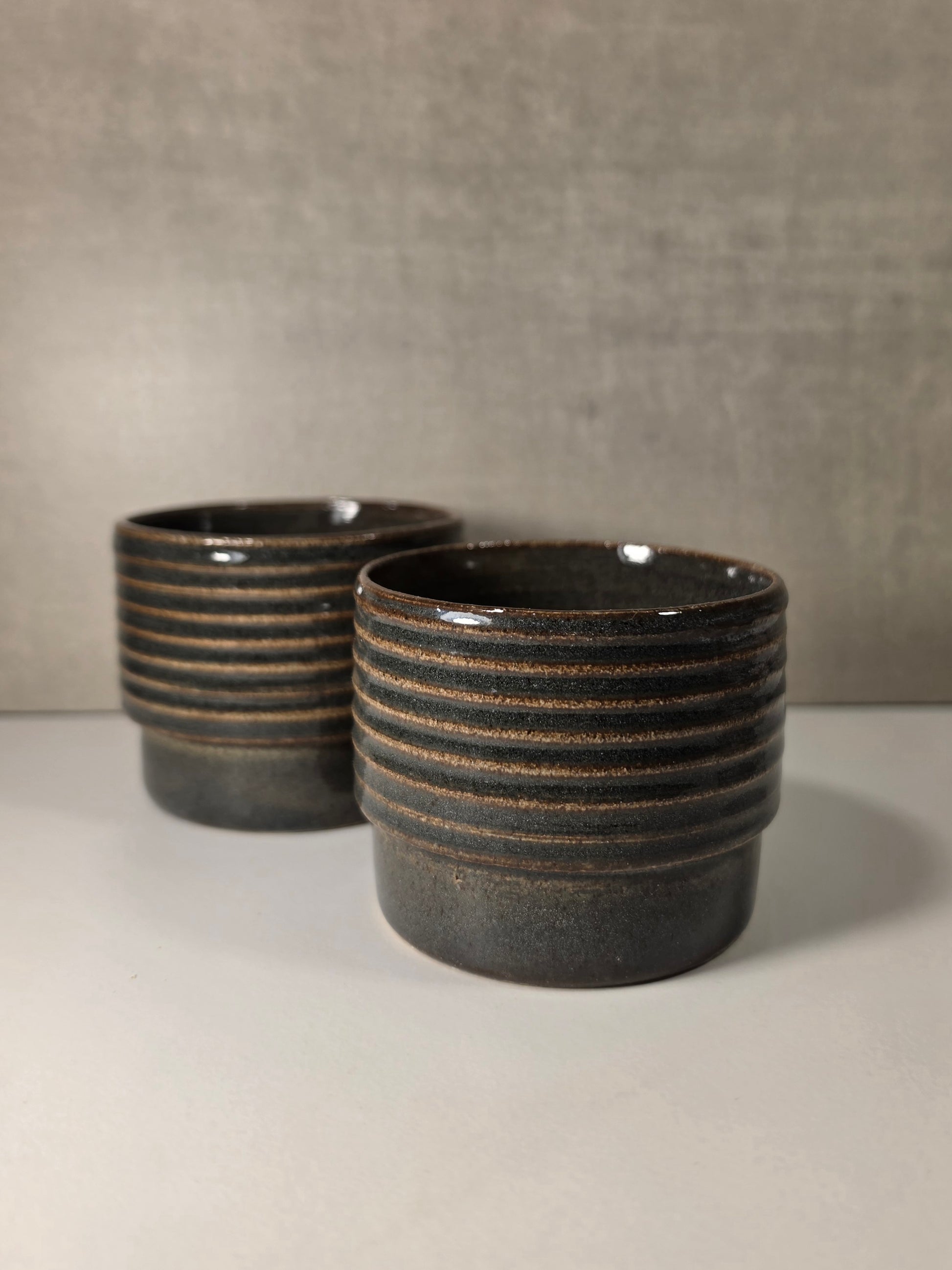 Two dark green glazed ceramic tumblers with horizontal ridges, displayed on a light gray surface against a textured gray background.