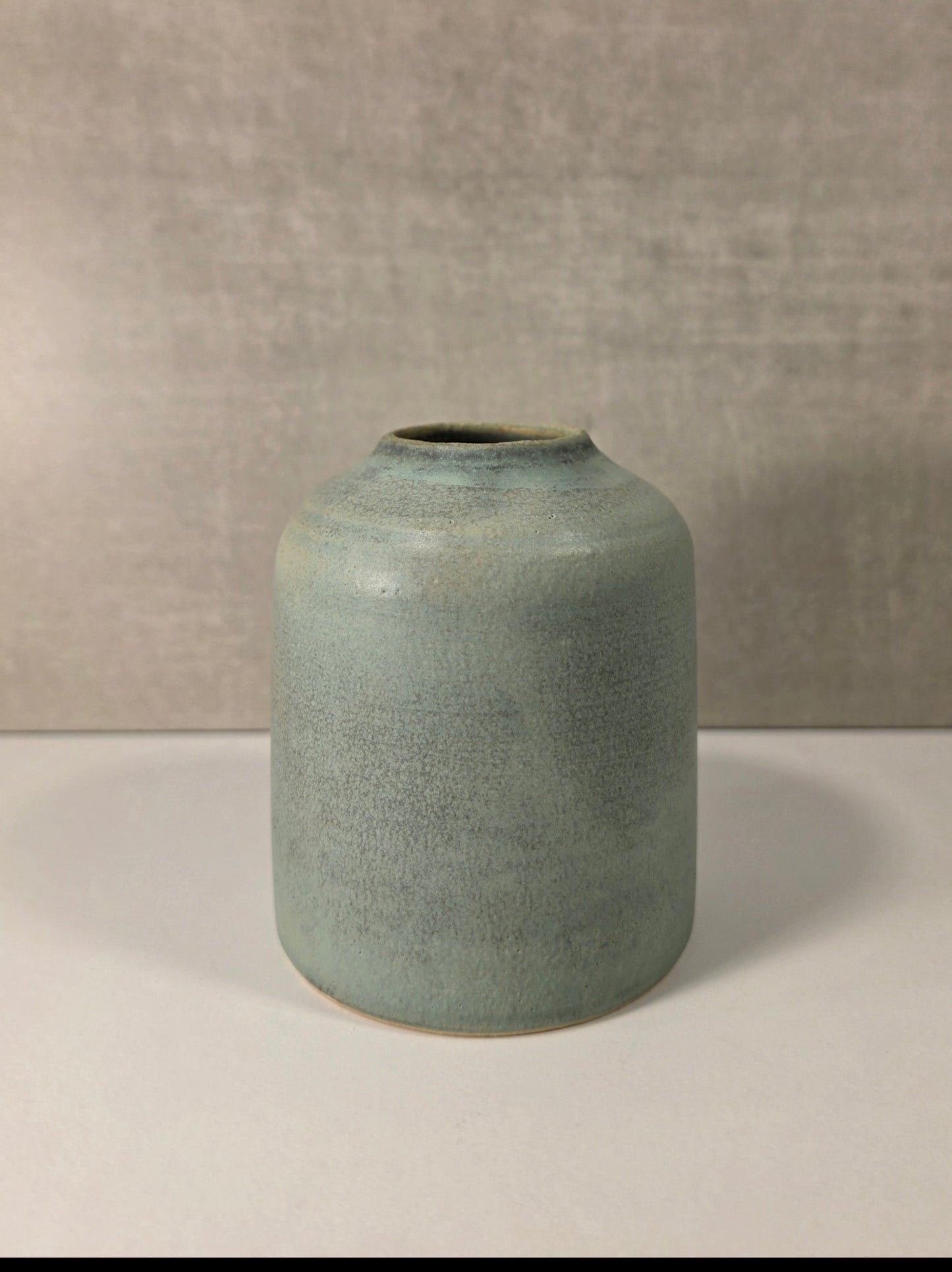 A small cylindrical ceramic vase with a wide mouth, featuring a textured sage green glaze and a natural clay foot, set against a neutral gray textured background.