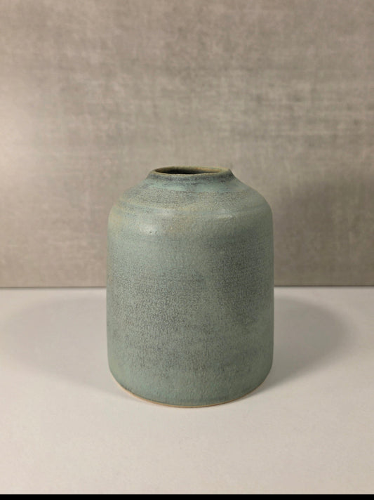 A small cylindrical ceramic vase with a wide mouth, featuring a textured sage green glaze and a natural clay foot, set against a neutral gray textured background.