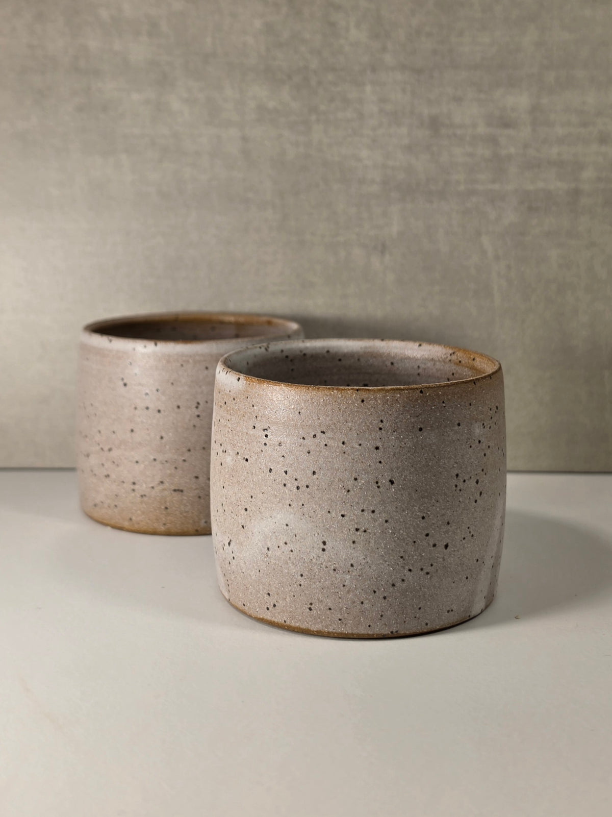 Two handmade ceramic planters with a speckled beige and gray glaze, displayed on a light surface against a textured gray background.