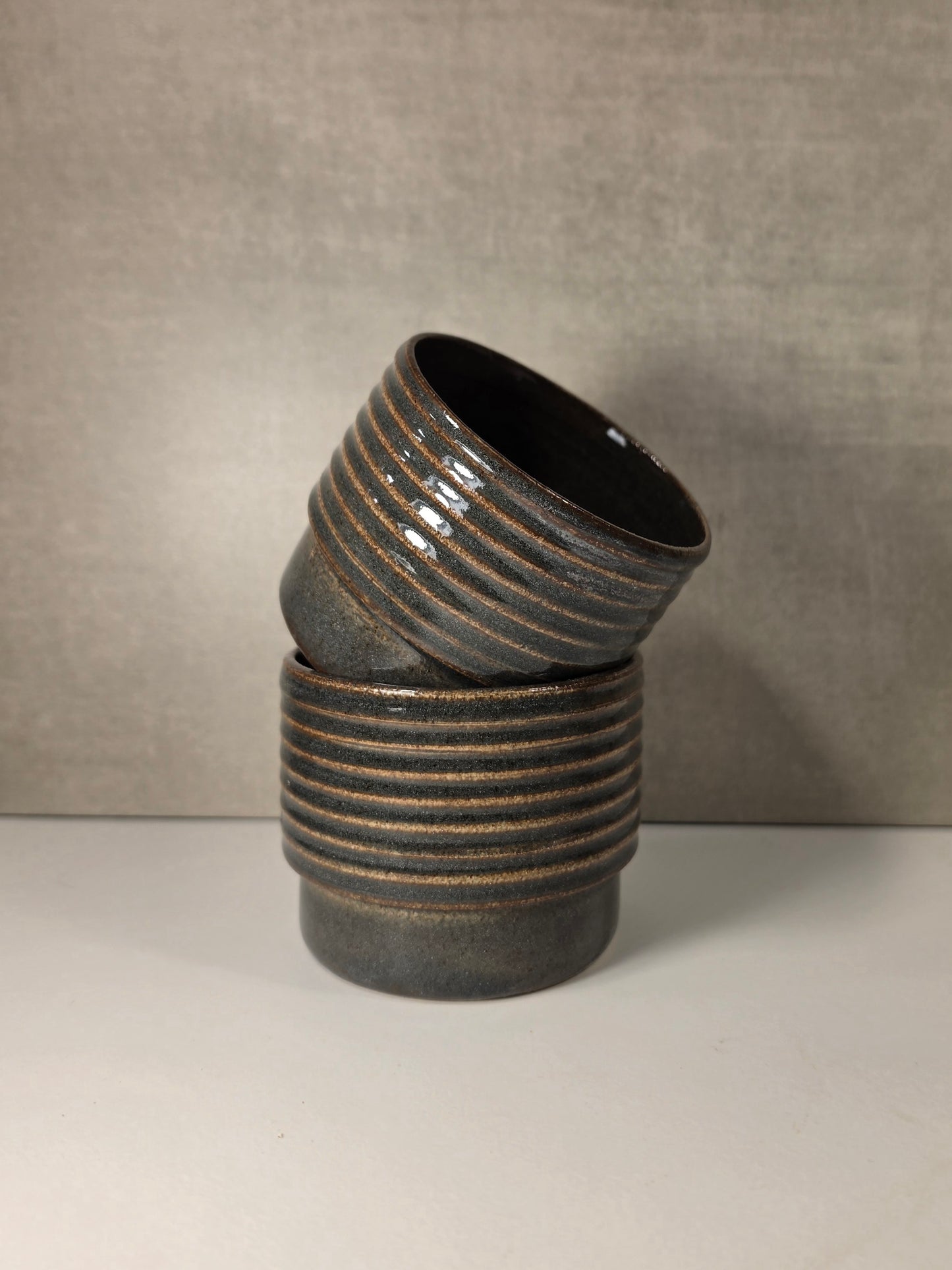 Two stacked ceramic cups with a dark greenish blue glaze, featuring horizontal ridges and a glossy finish, set against a neutral gray background.