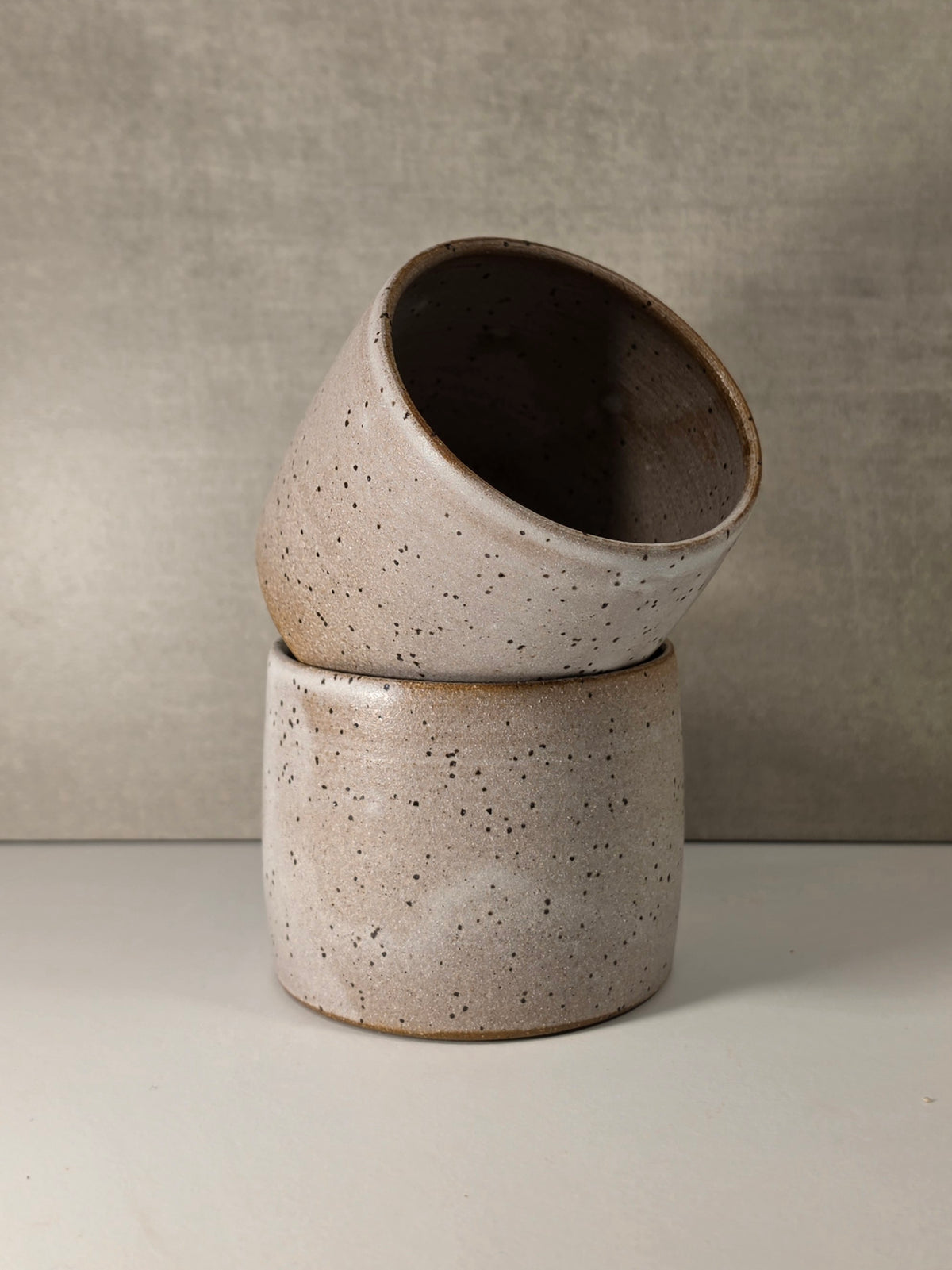 Two stacked ceramic planters with a speckled beige and brown glaze, set against a neutral textured gray background.