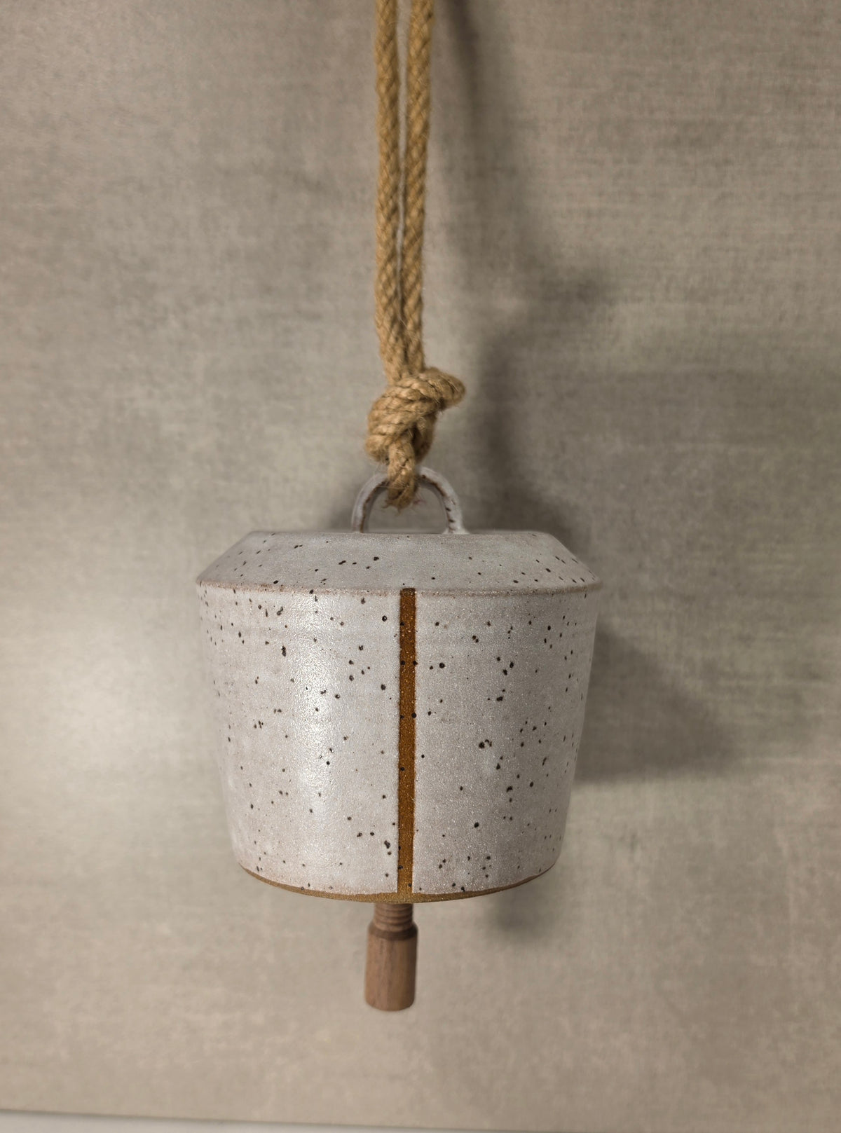 A small speckled ceramic bell with a natural wood spout and a hanging rope, set against a textured beige background.
