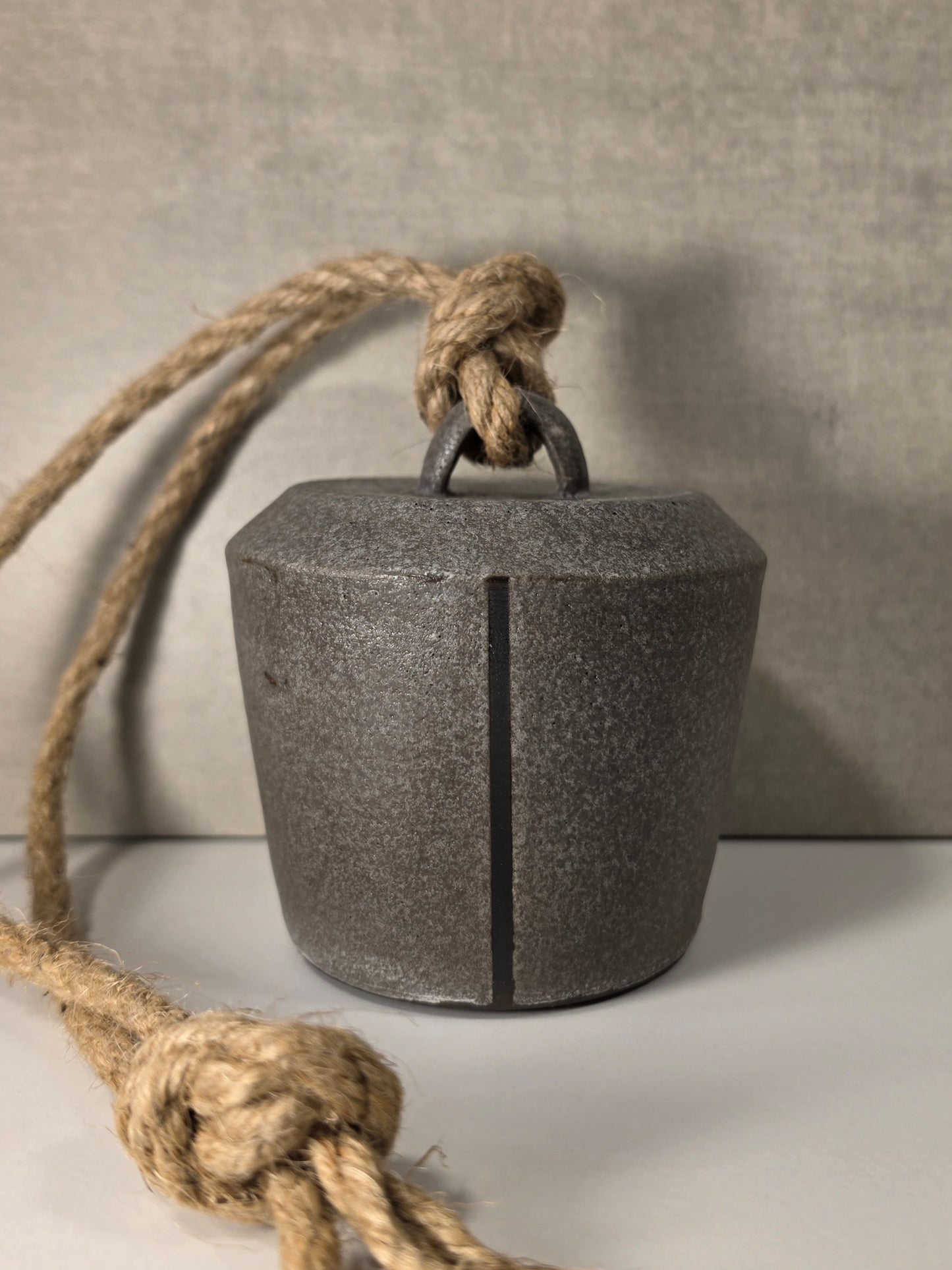 A dark ceramic bell glazed to look like cast iron bell with a natural jute rope hanger, shown on a neutral gray background.