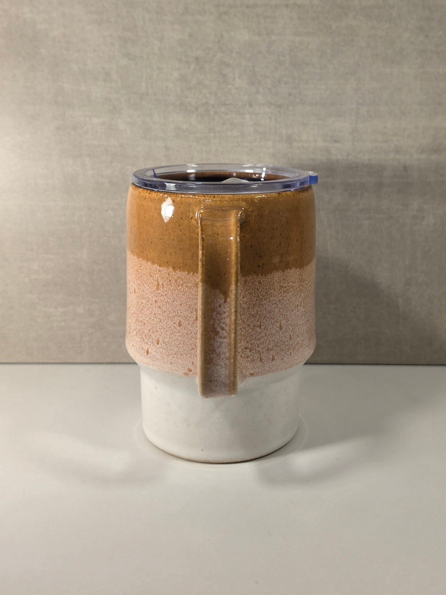 A handmade ceramic mug with a three-tiered glaze design, featuring a dark brown top section, a speckled pinkish-brown middle section with a vertical handle, and a white bottom section. It is topped with a clear lid that has a blue rim, set against a neutral gray textured background.