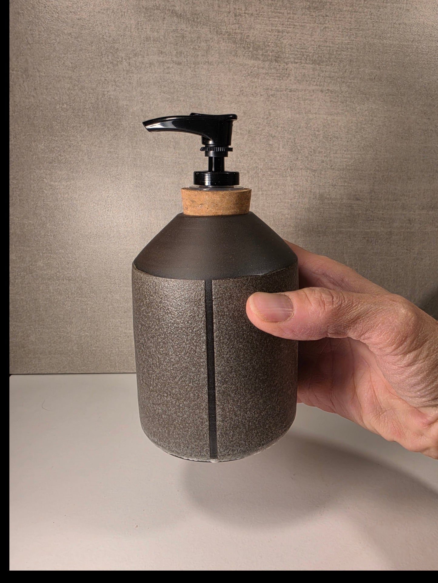 A hand holding a matte dark gray ceramic soap dispenser with a black pump and a light wood collar, set against a textured gray wall and white surface.
