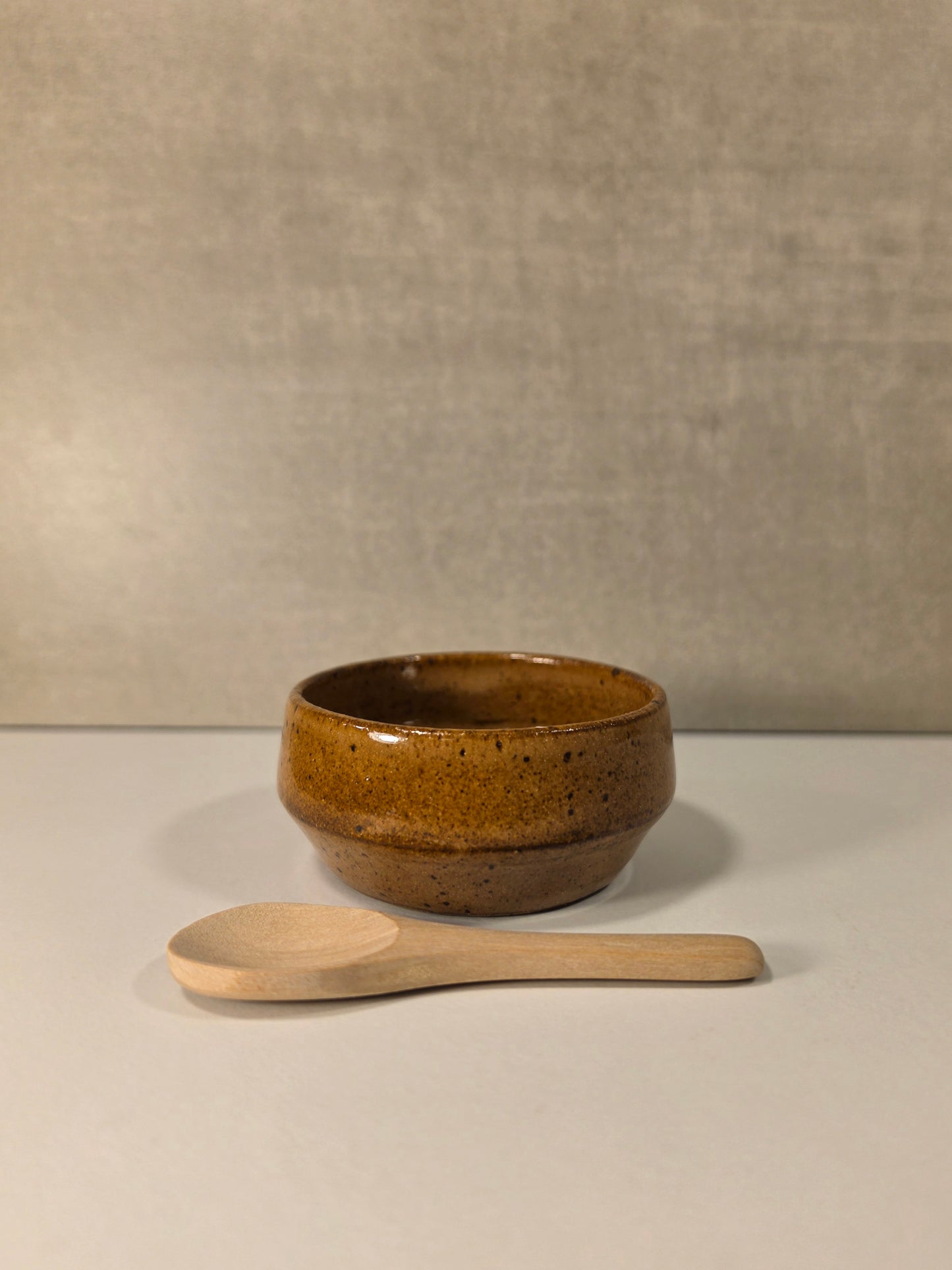 A small round brown ceramic bowl with a speckled glaze sits on a light surface, with a light wood spoon placed in front against a neutral textured backdrop.