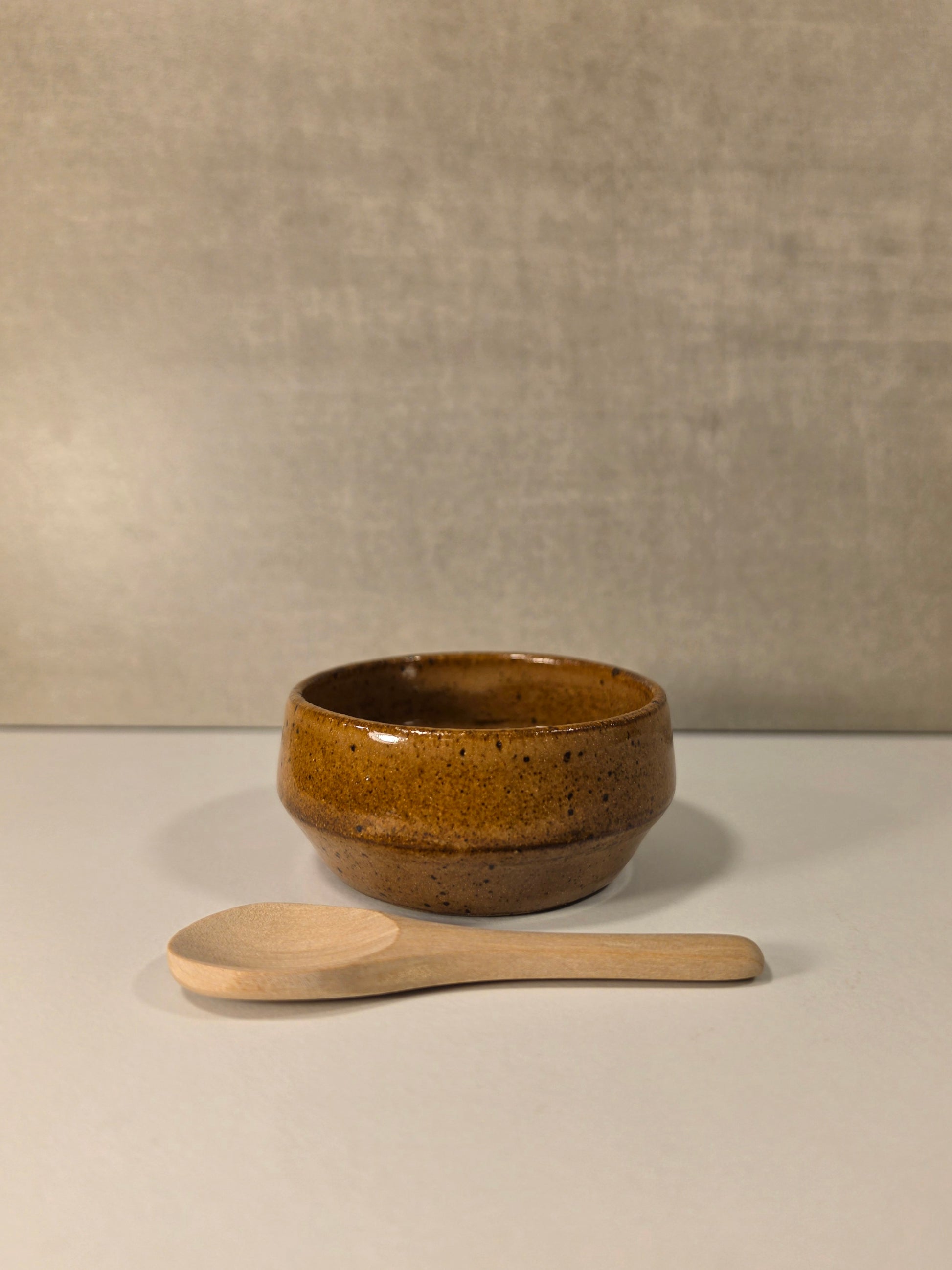 A small round brown ceramic bowl with a speckled glaze sits on a light surface, with a light wood spoon placed in front against a neutral textured backdrop.