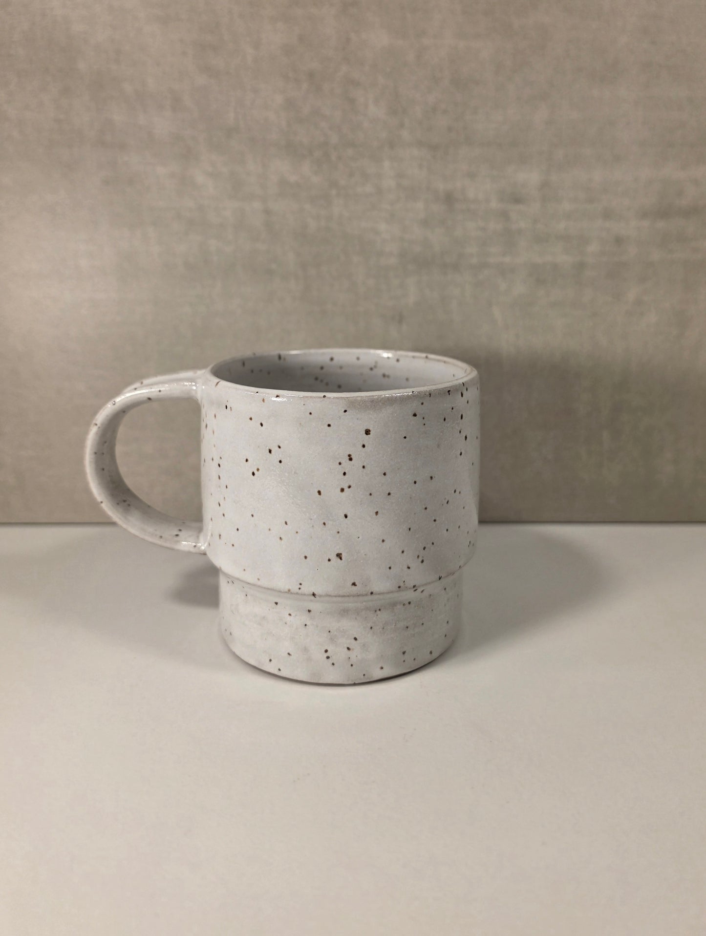 A handmade ceramic mug with a speckled white glaze and dark brown speckles, set against a neutral textured gray background.