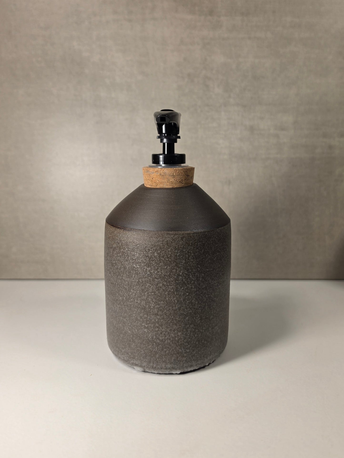 A dark gray textured ceramic dispenser bottle with a cork neck and a black pump, set against a neutral gray background.