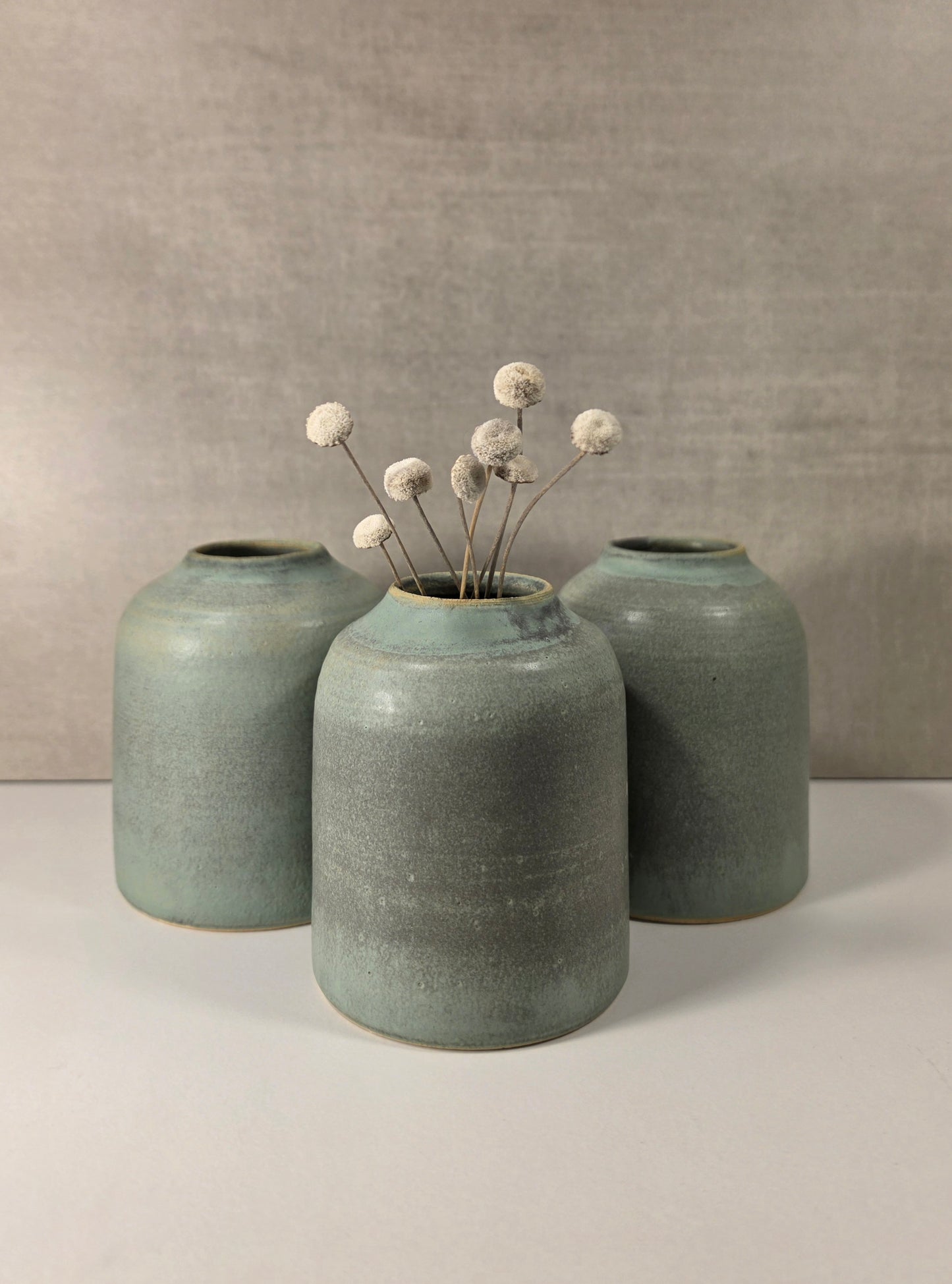 Three matte ceramic vases in a muted sage green glaze, with the center vase holding a small dried floral arrangement, set against a neutral gray textured background.