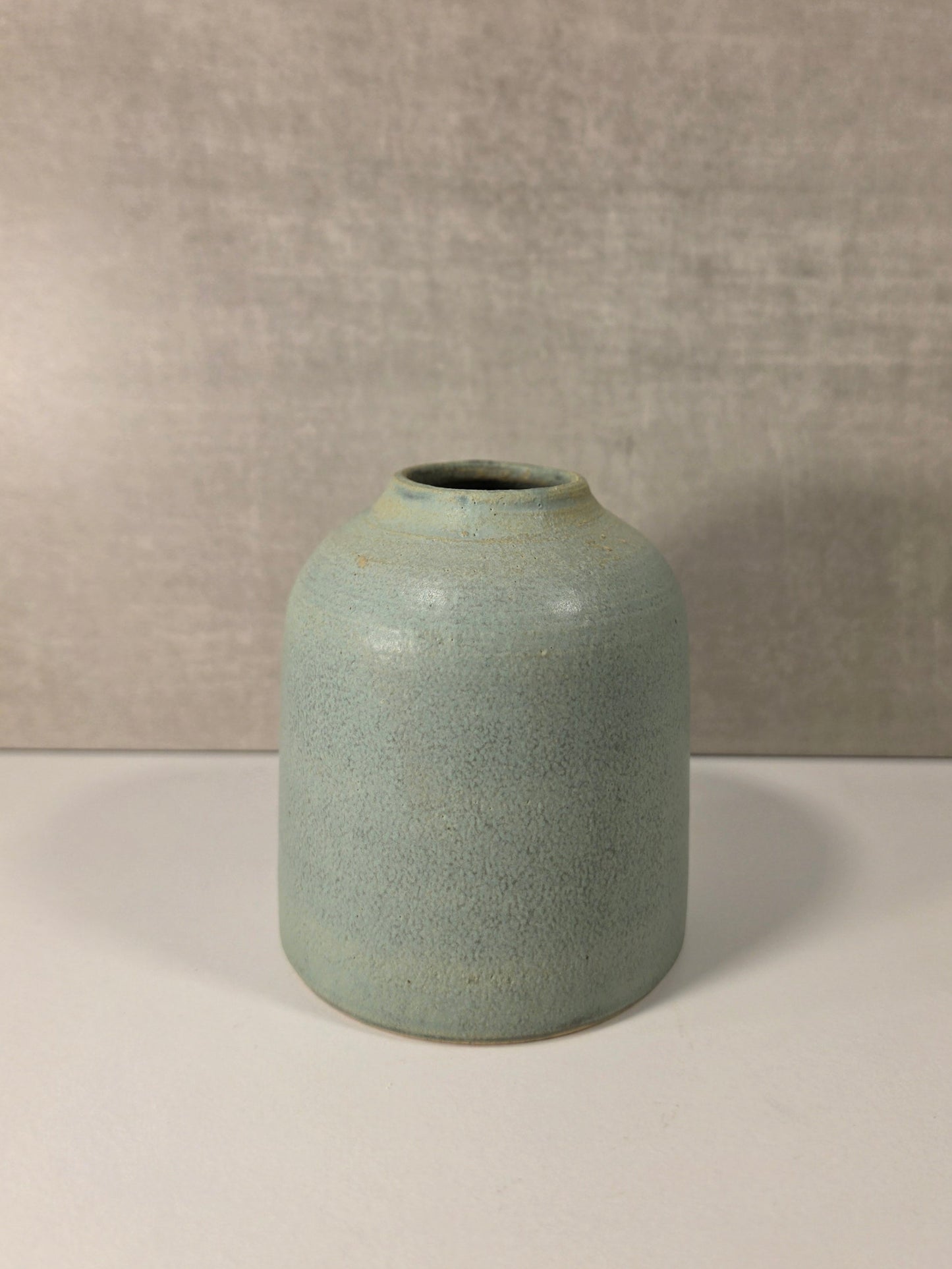 A small ceramic vase with a rounded cylindrical shape and a short opening, featuring a speckled light blue-green glaze. It is placed on a white surface against a textured gray wall.