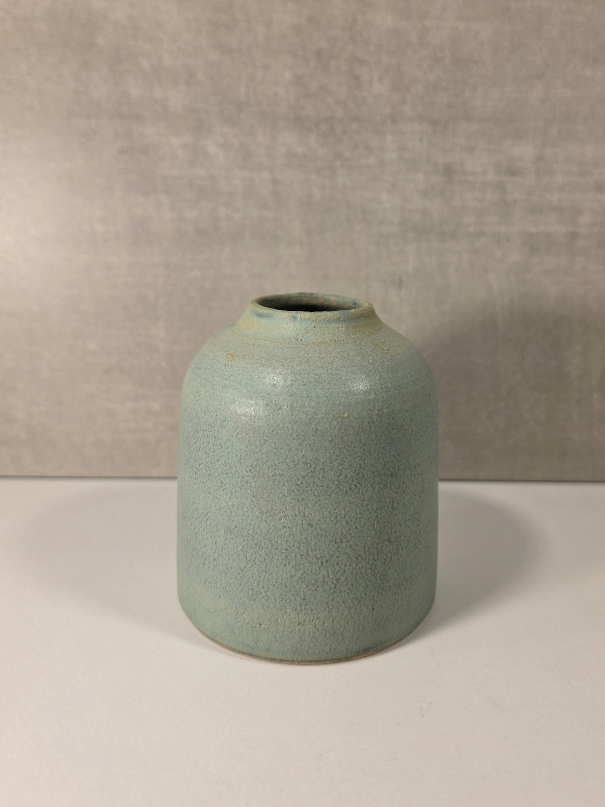 A small ceramic vase with a rounded cylindrical shape and a short opening, featuring a speckled light blue-green glaze. It is placed on a white surface against a textured gray wall.