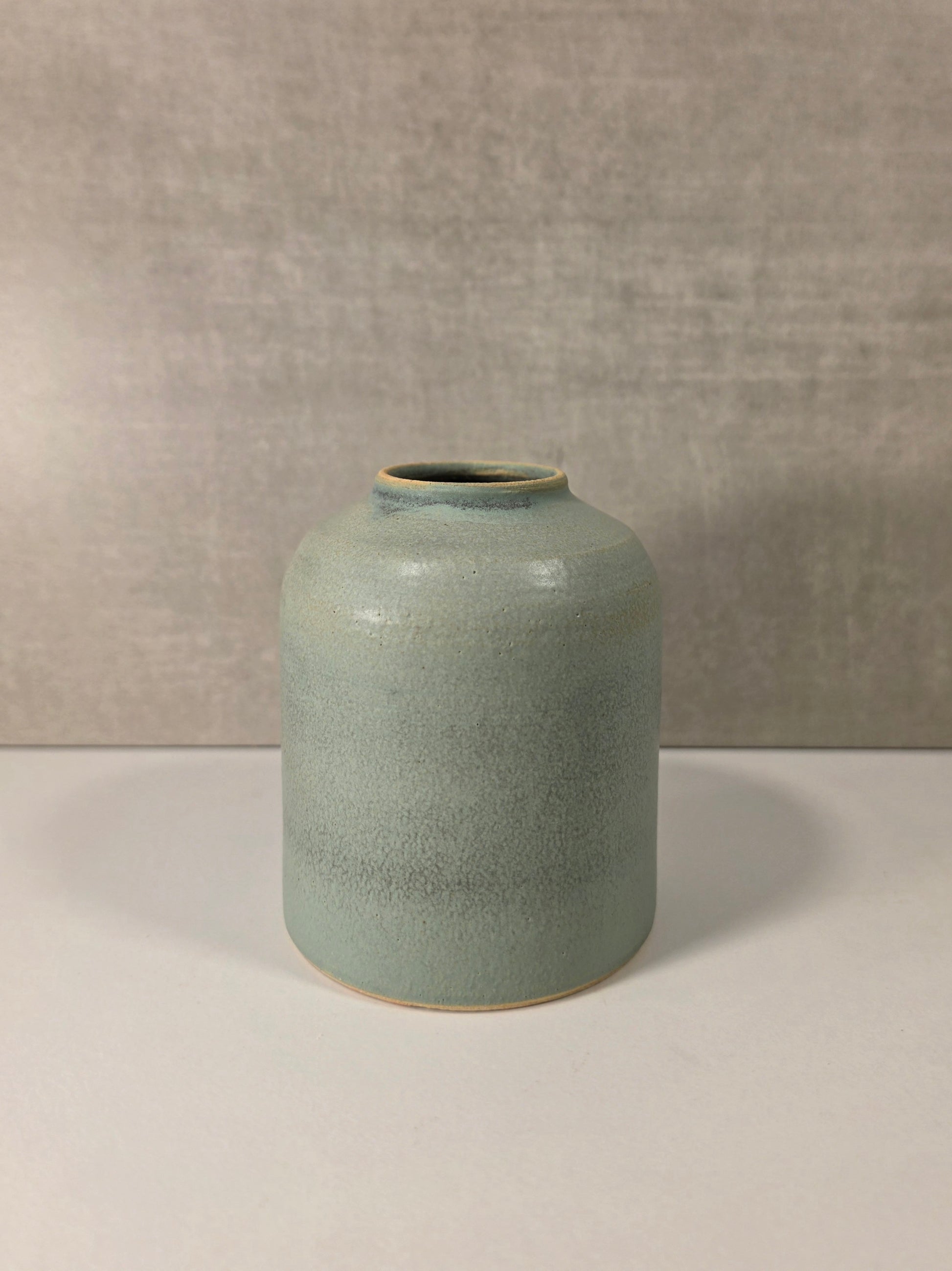 A small ceramic vase with a rounded body and short neck, featuring a speckled sage green glaze with a subtle blue undertone and a light beige unglazed base, set against a neutral gray textured background.