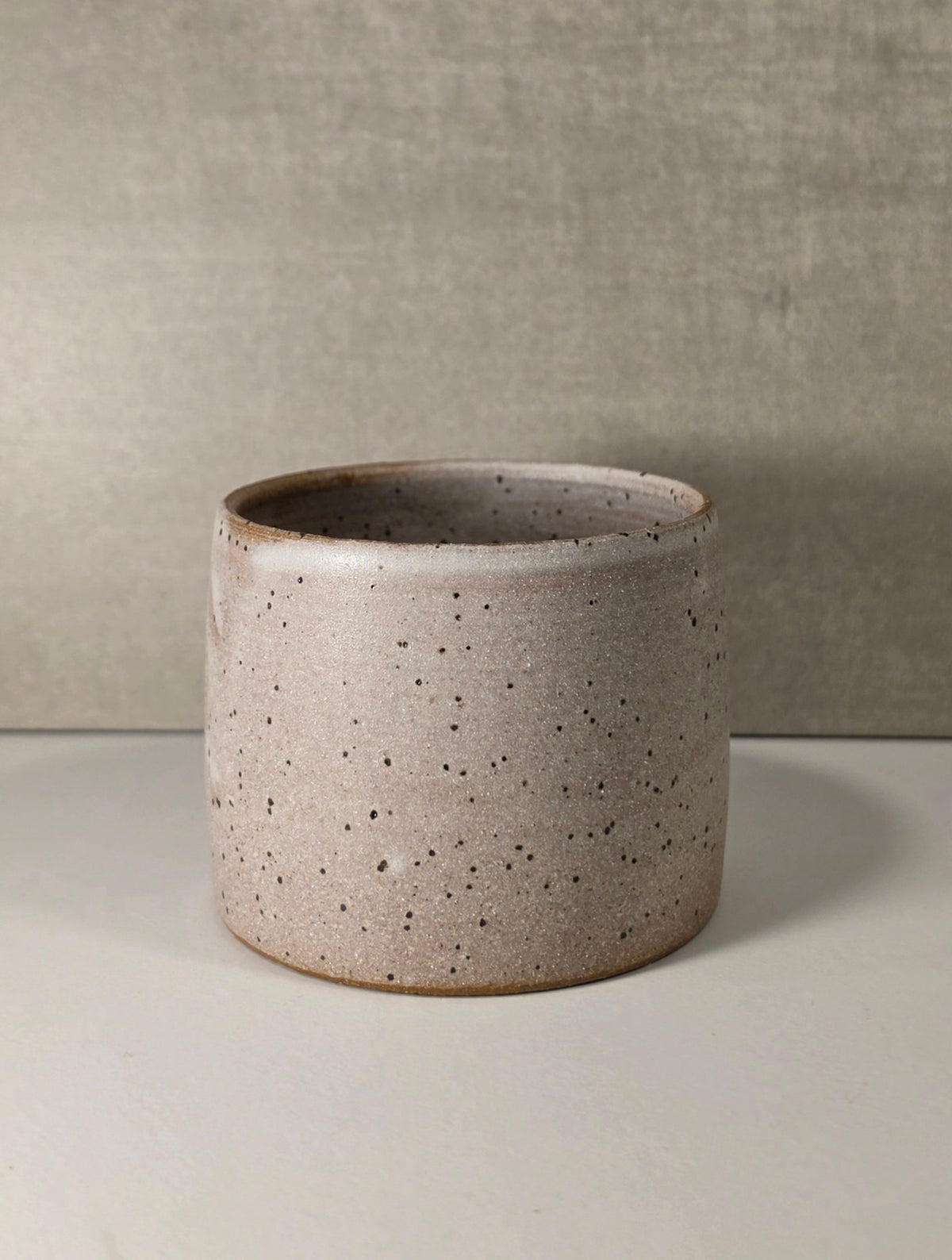 A cylindrical ceramic cappuccino mug or cortado cup with a speckled beige and brown glaze, set against a neutral textured gray background.