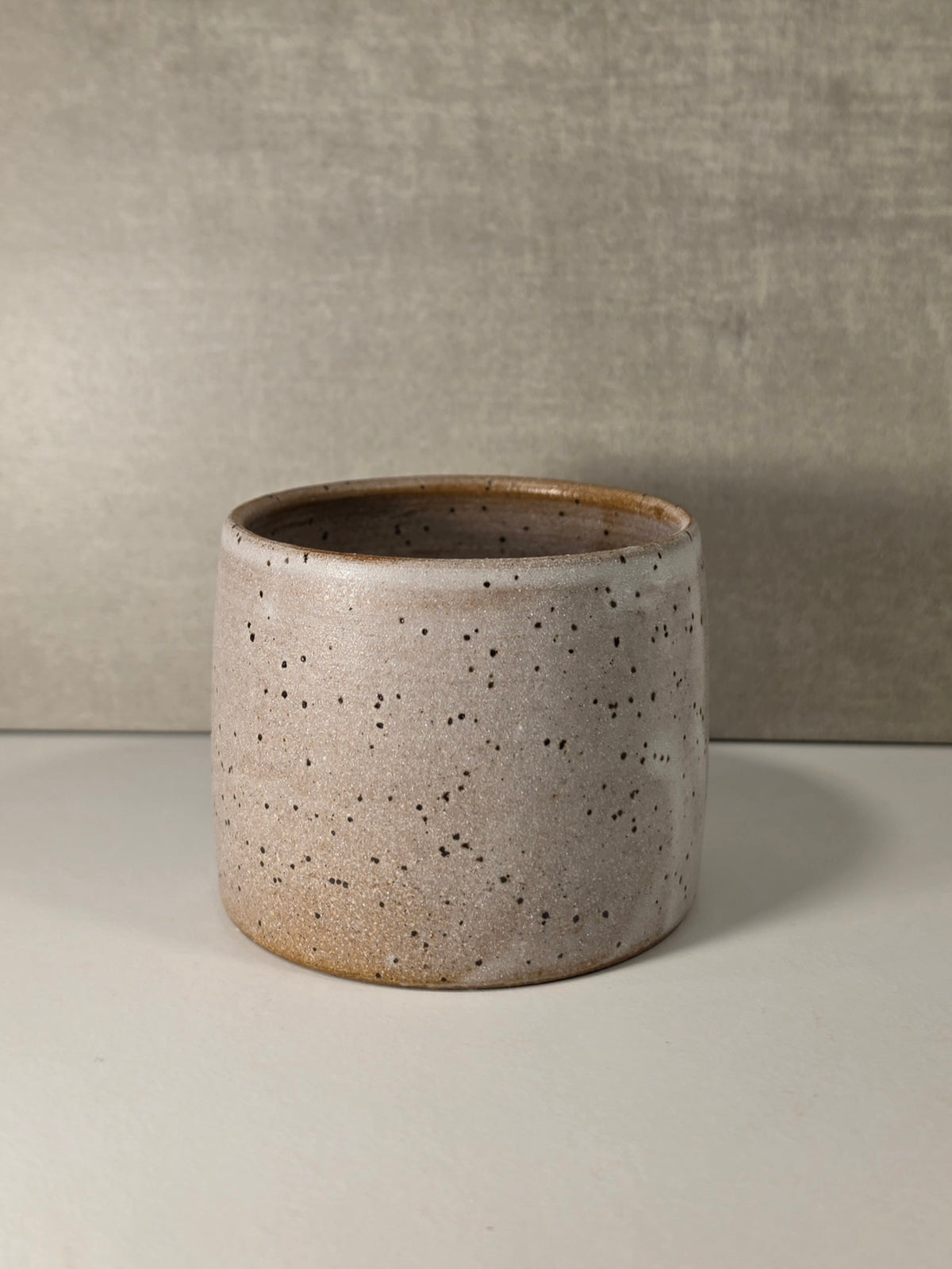 A cylindrical ceramic planter with a speckled beige and brown glaze, set against a neutral gray textured background.