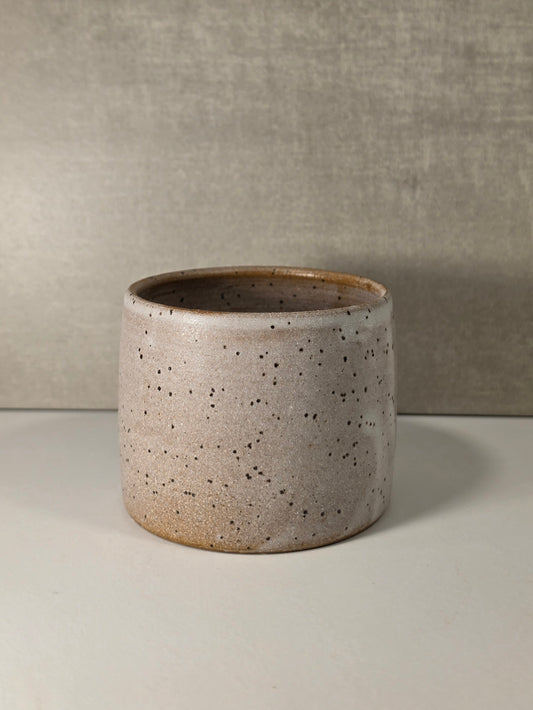 A cylindrical ceramic planter with a speckled beige and brown glaze, set against a neutral gray textured background.