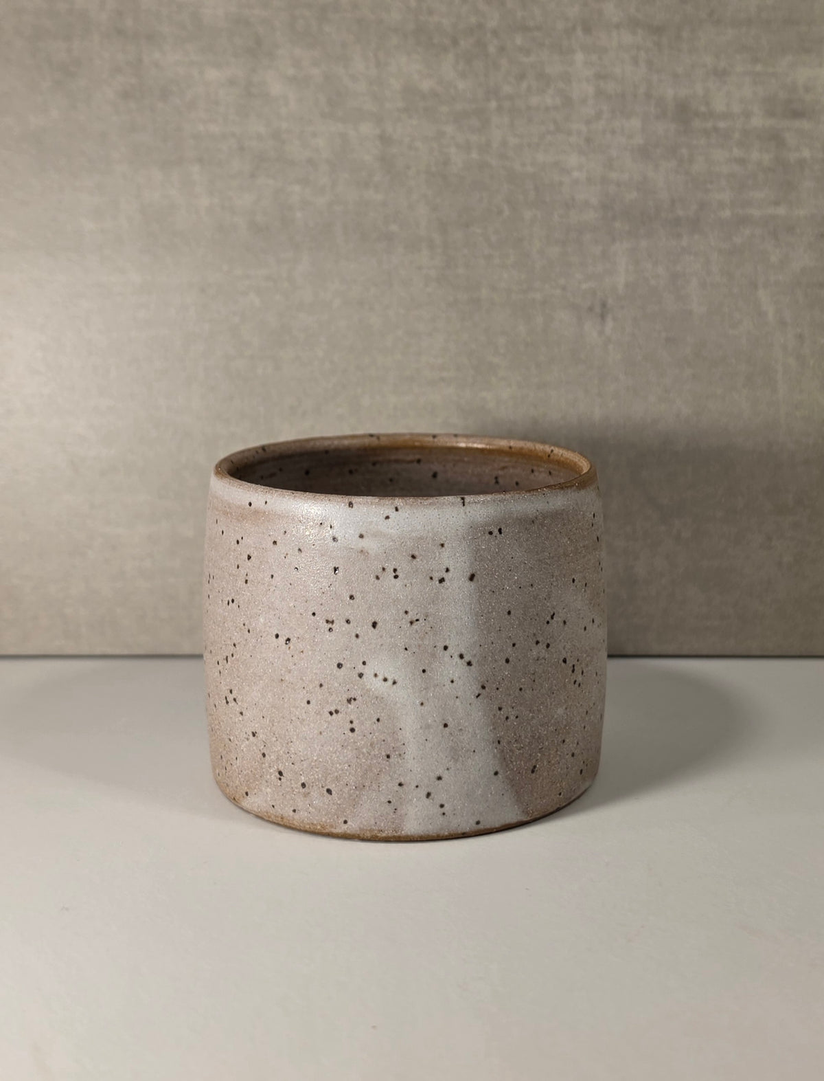 A handmade ceramic cup with a speckled beige and white glaze and a brown rim, set against a neutral gray textured background.