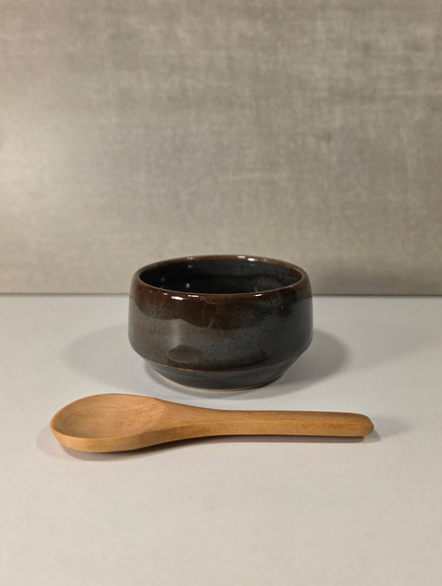 A small dark glazed ceramic bowl with a wooden spoon placed in front, set against a neutral gray textured background.