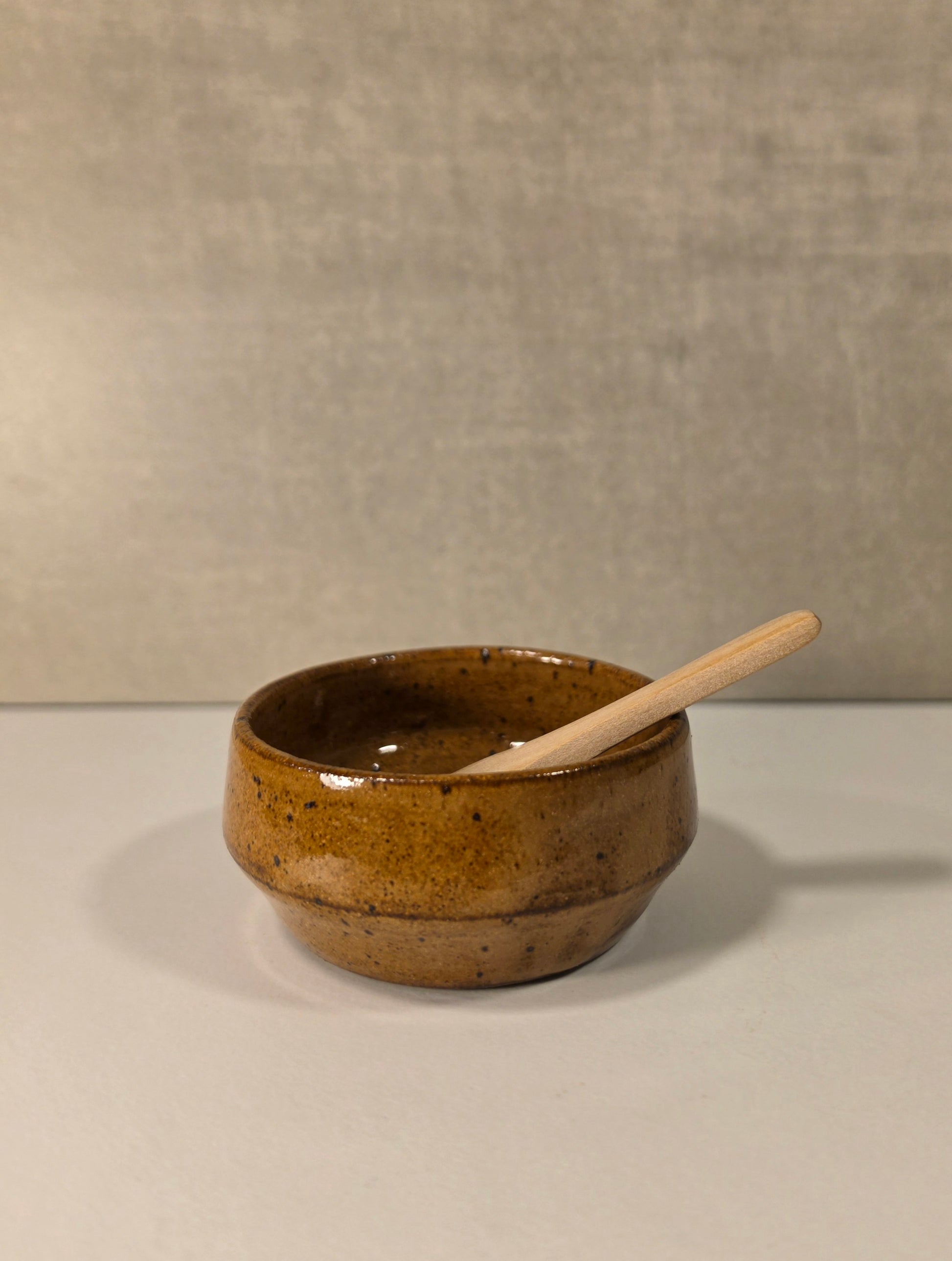 A small handmade ceramic bowl with a speckled brown and amber glaze, containing a light-colored wooden spoon, set against a neutral textured gray background.