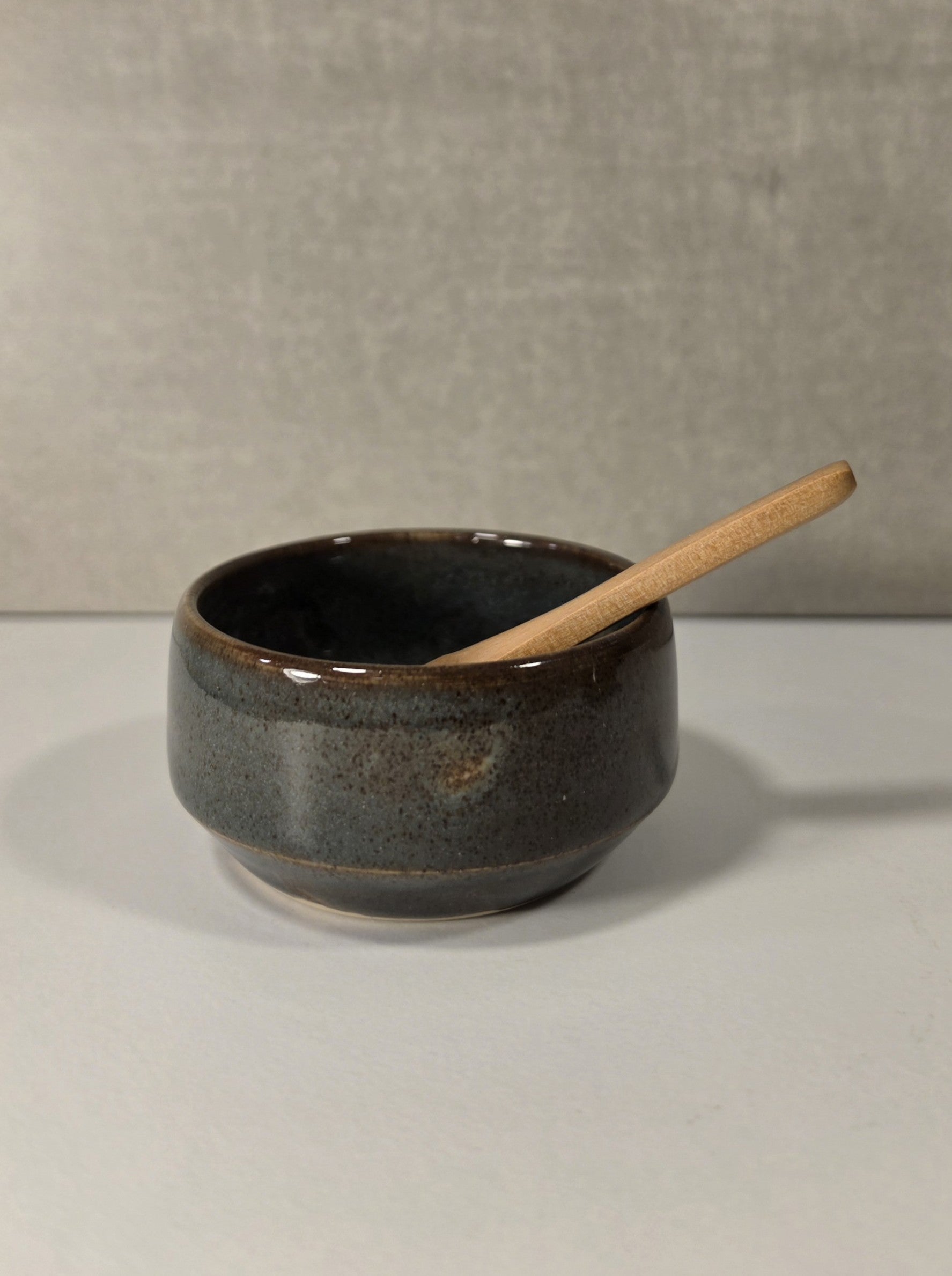 A small handmade ceramic bowl with a dark brown and black speckled glaze, containing a light natural wood spoon, set against a neutral gray textured background.