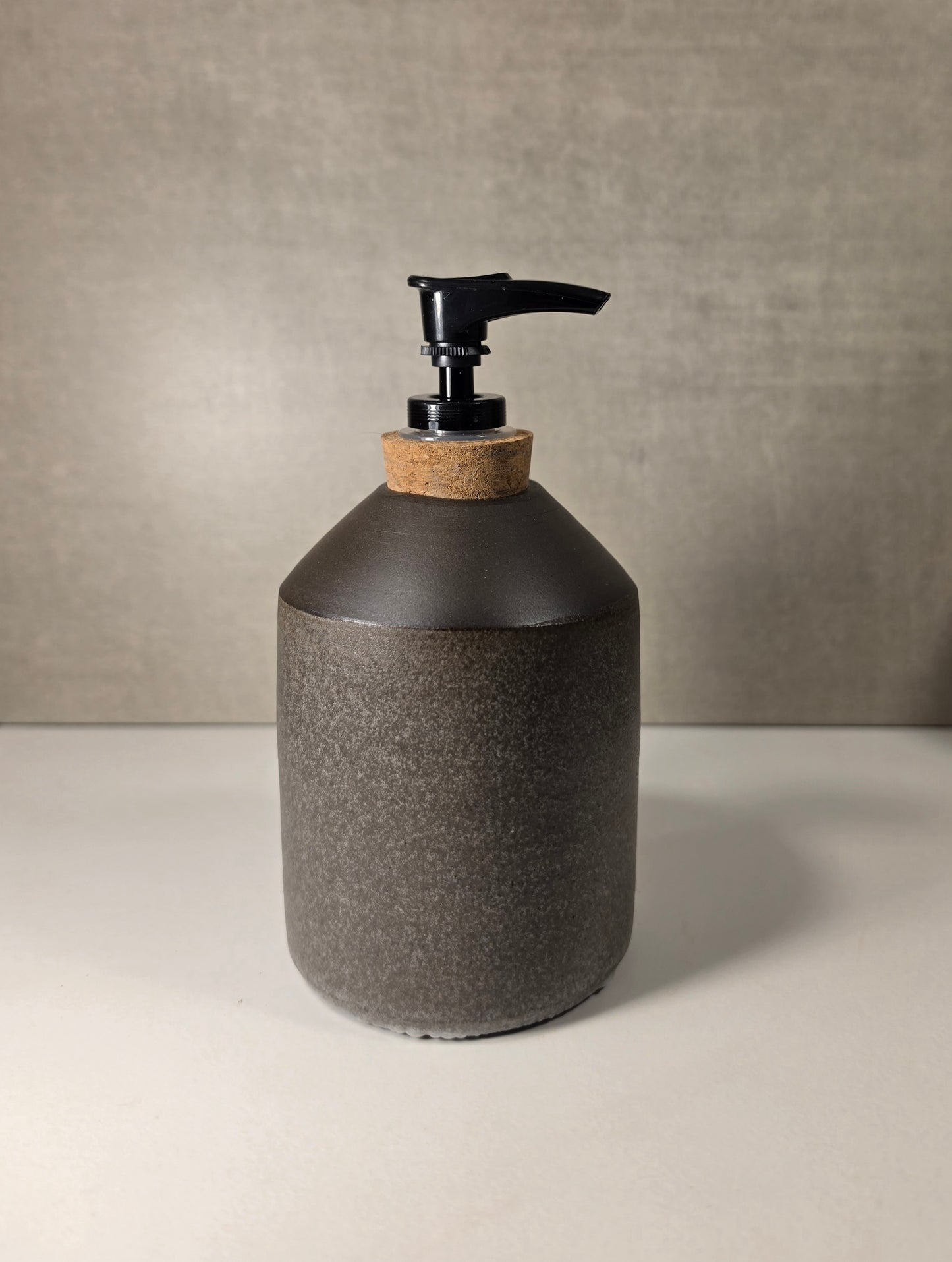 A dark gray textured ceramic soap dispenser with a black pump and a natural cork collar, set against a neutral gray background.