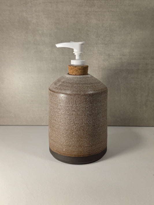 A ceramic pump dispenser bottle with a speckled brown and beige glaze, a cork neck, and a white plastic pump, set against a textured gray background.