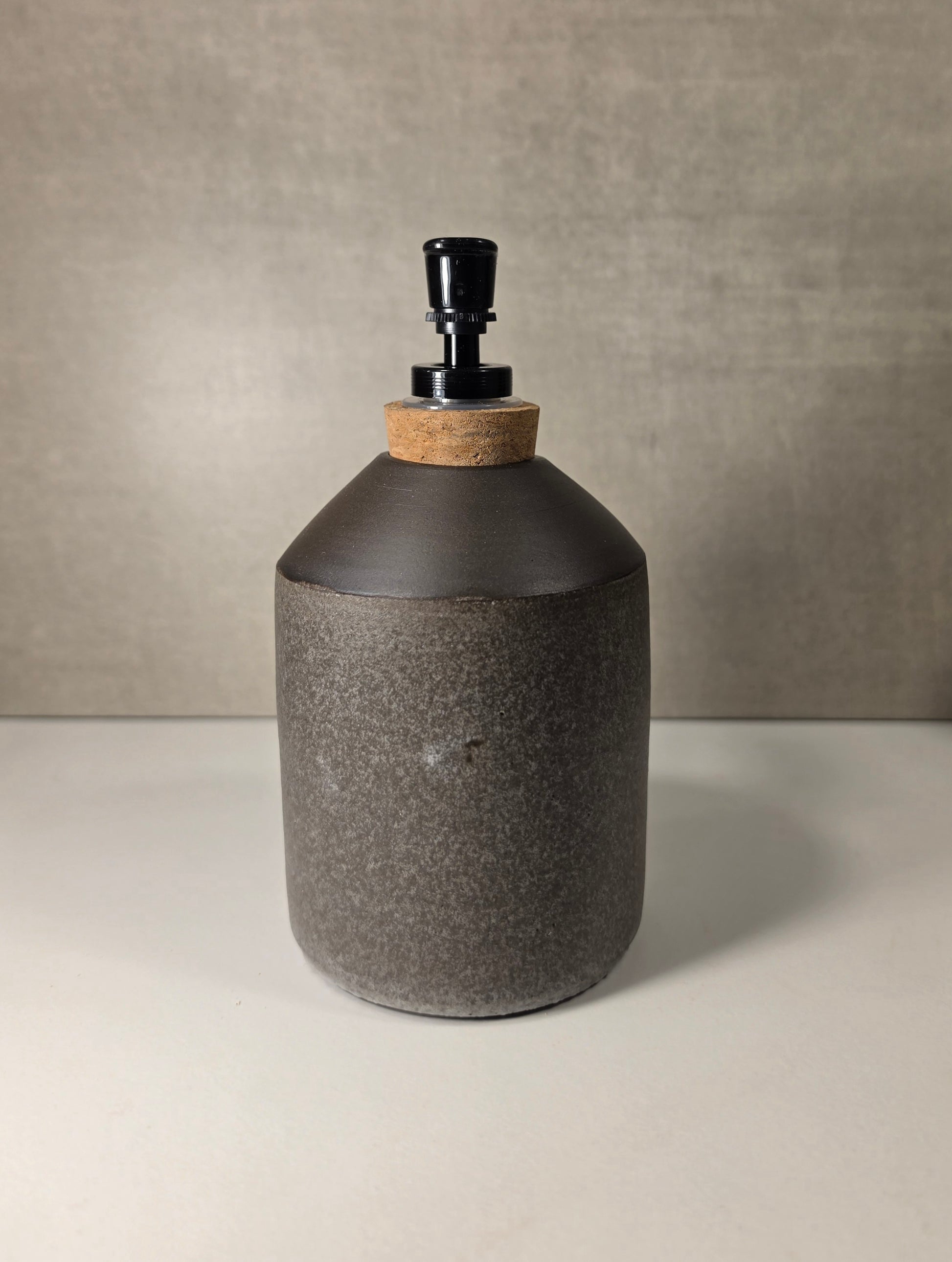 A dark gray textured ceramic bottle with a matte charcoal top, a natural cork neck, and a black pump dispenser, set against a neutral gray background.