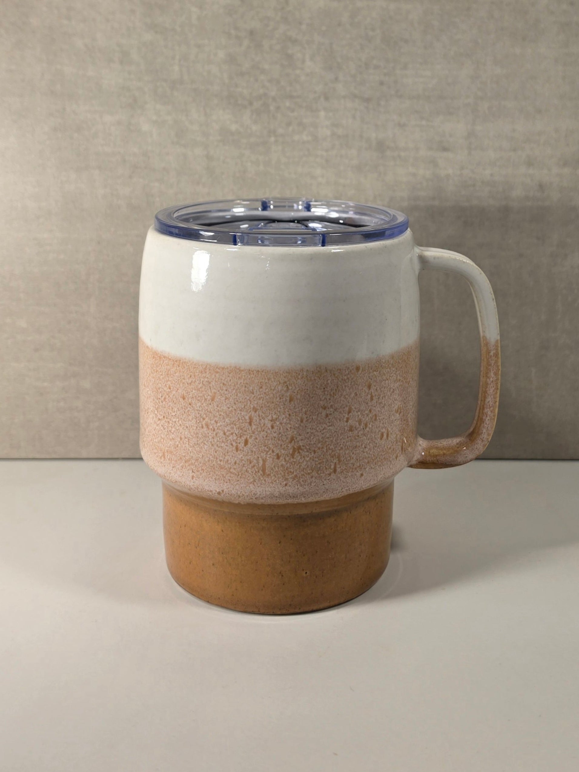 A ceramic travel mug with a three-tiered color-blocked design in white, speckled beige, and earthy brown, featuring a clear plastic lid with a blue rim and a side handle, set against a neutral textured gray background.