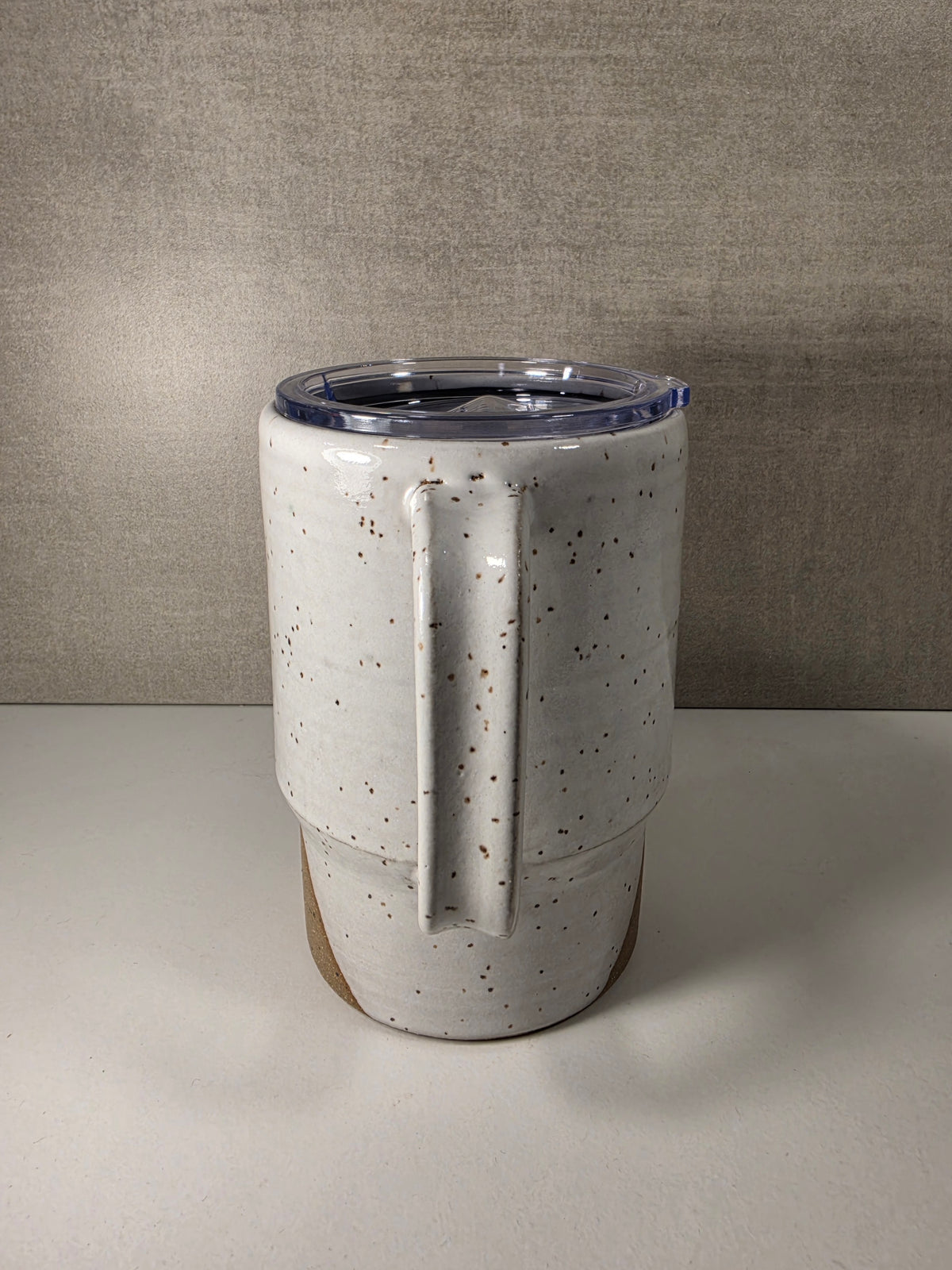 A ceramic tumbler with a textured speckled glaze in off-white and brown, featuring a large integrated handle, a clear plastic lid, and a visible wood-toned base, set against a neutral gray background.