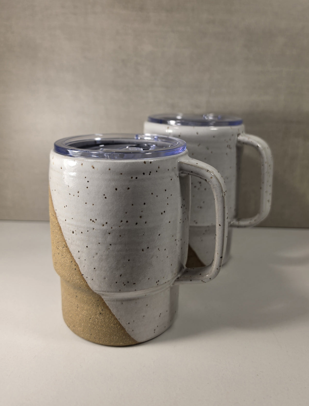 Two ceramic travel mugs with handles and clear lids, one in the foreground and one blurred in the background. The mugs have a speckled off-white glaze with a diagonal tan section on the lower half, set against a neutral textured gray background.