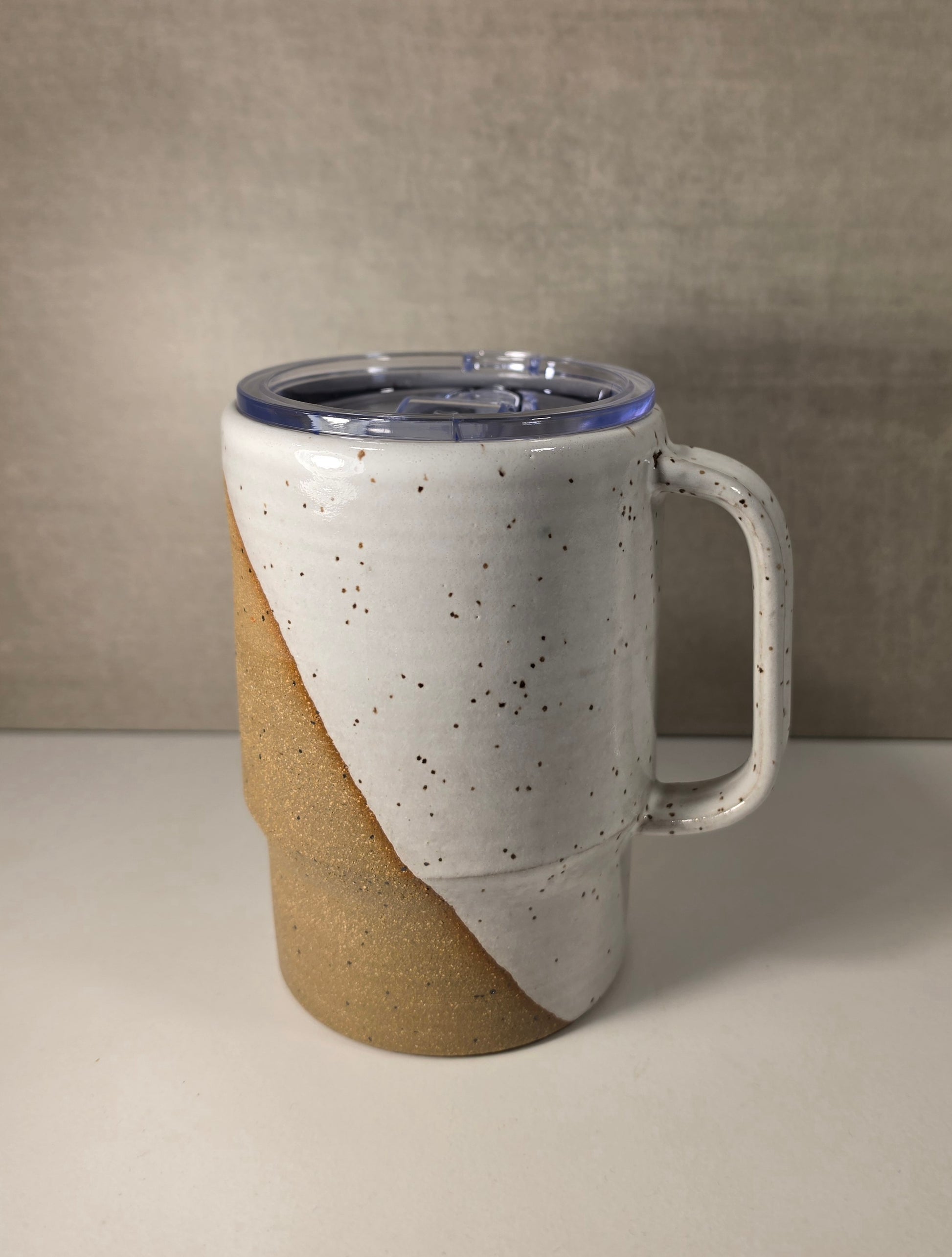 A ceramic travel mug with a handle, featuring a white speckled glaze on the upper half and a natural tan clay finish on the lower half, topped with a clear lid.