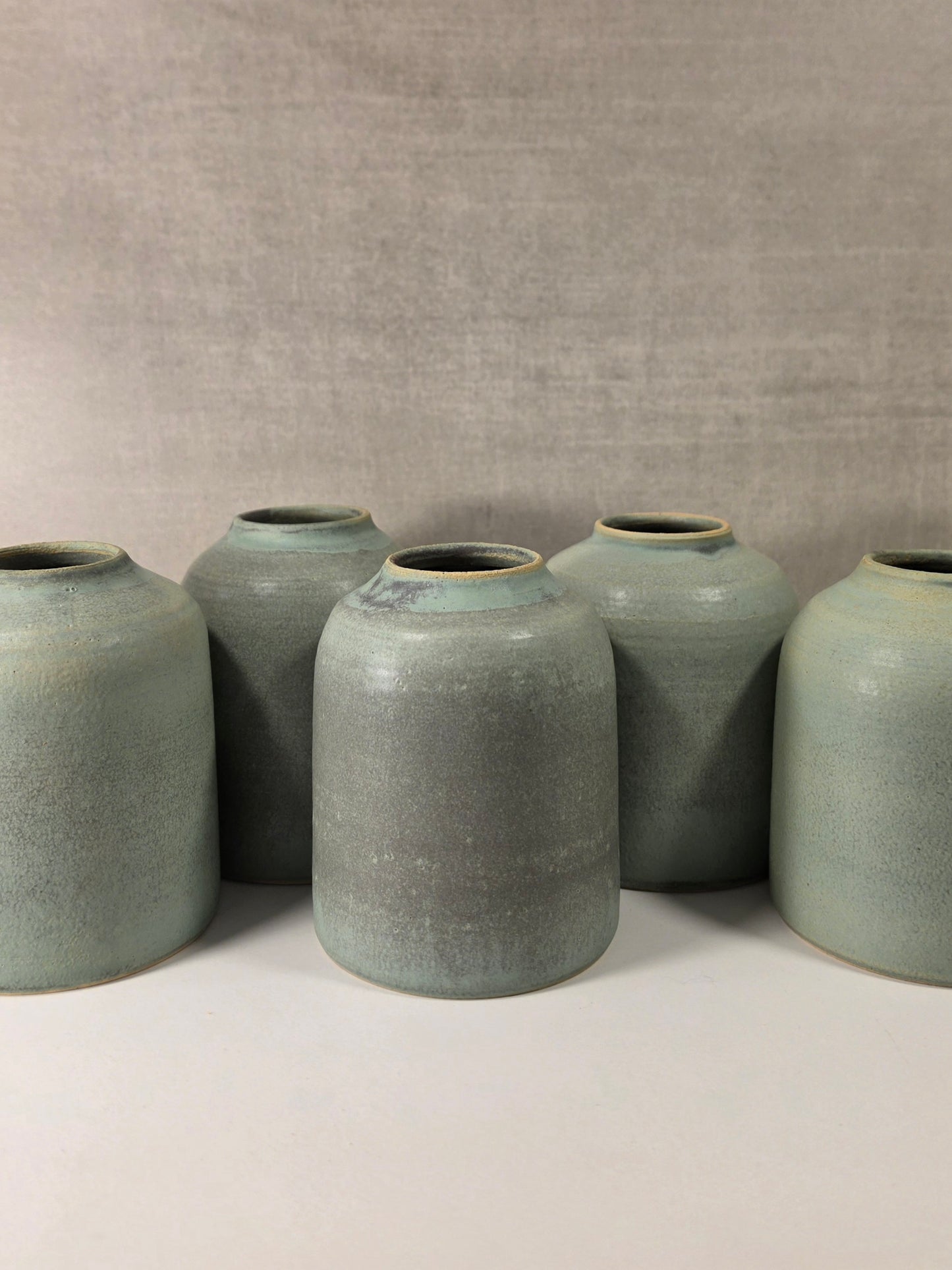 Five handmade ceramic vases with a textured sage green glaze, arranged in a row against a neutral gray fabric backdrop.
