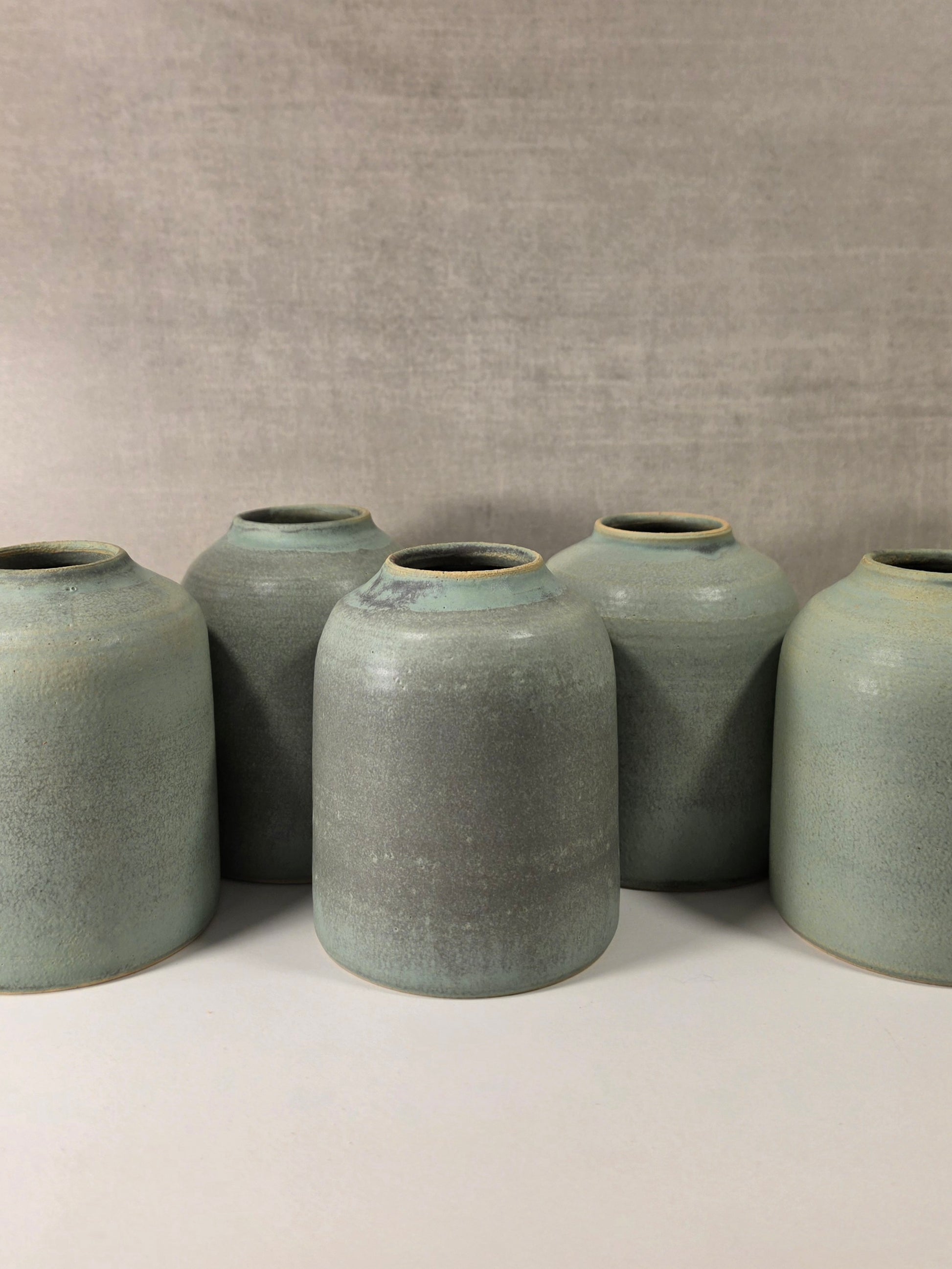 Five handmade ceramic vases with a textured sage green glaze, arranged in a row against a neutral gray fabric backdrop.