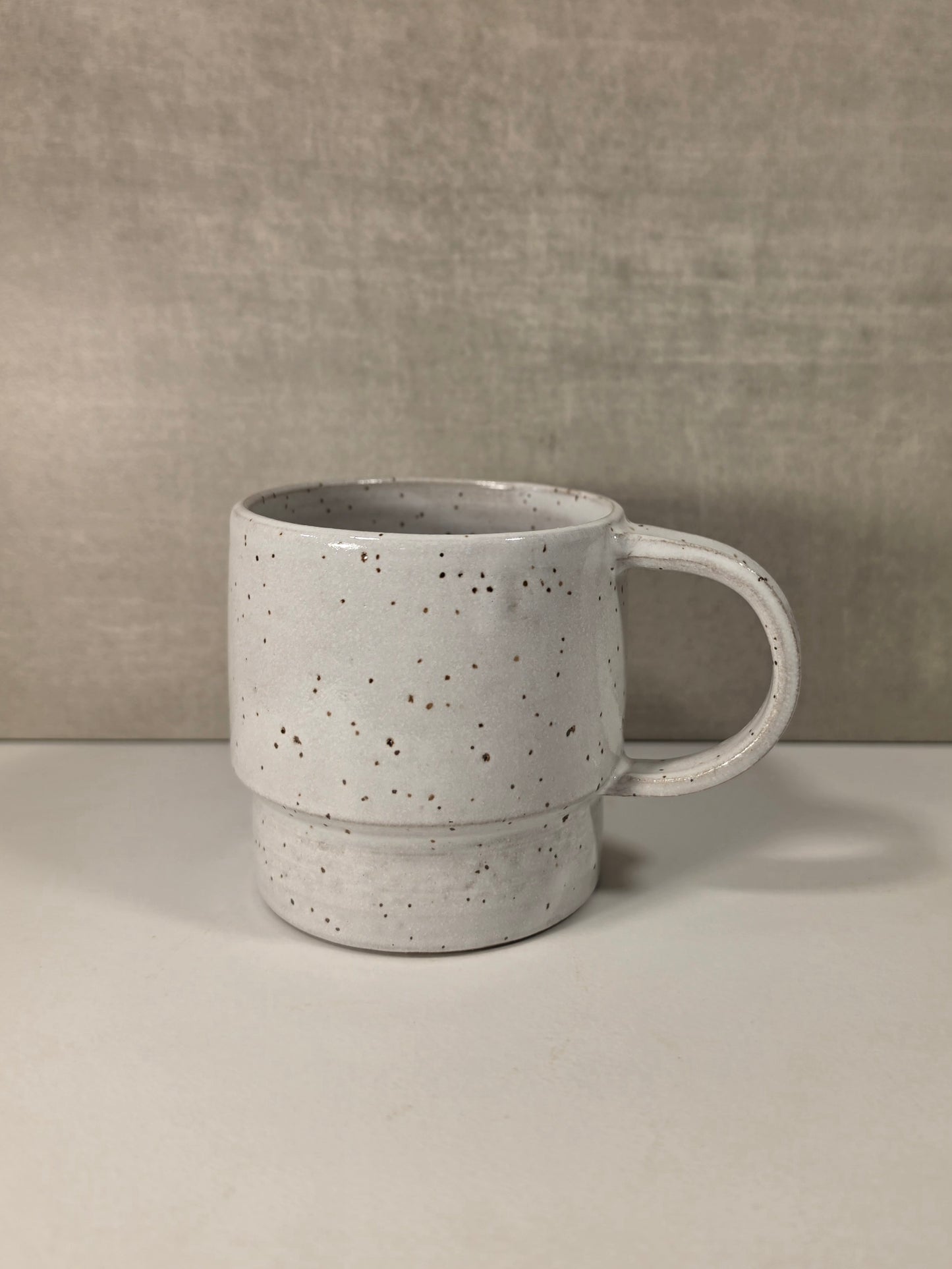 A handmade tiered ceramic mug with a textured, speckled white glaze and a thick handle, set against a neutral gray background.