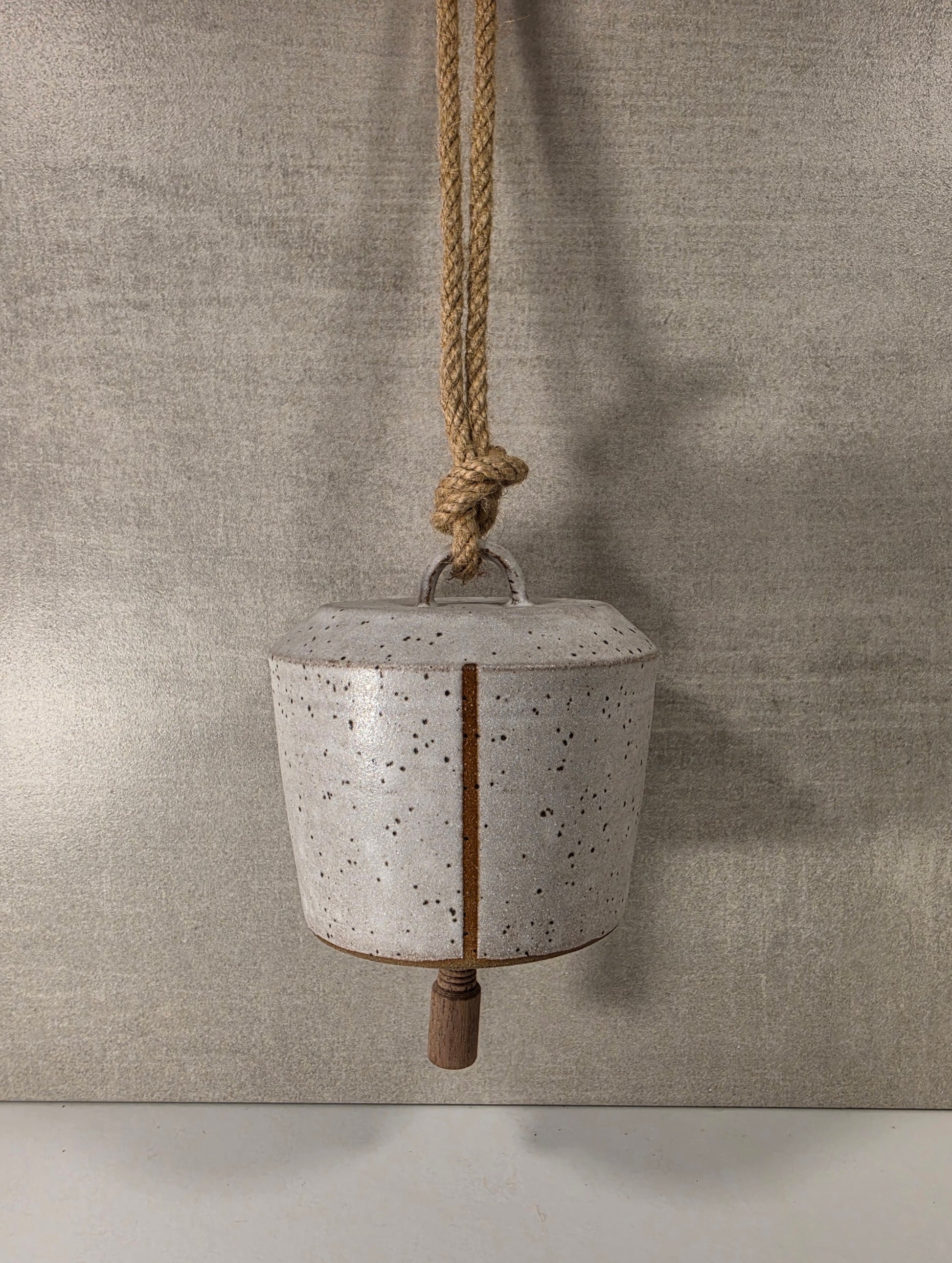 A rustic ceramic hanging bell with a speckled off-white glaze, a vertical brown crack, and a wooden mallet, suspended by a thick jute rope against a textured gray background.