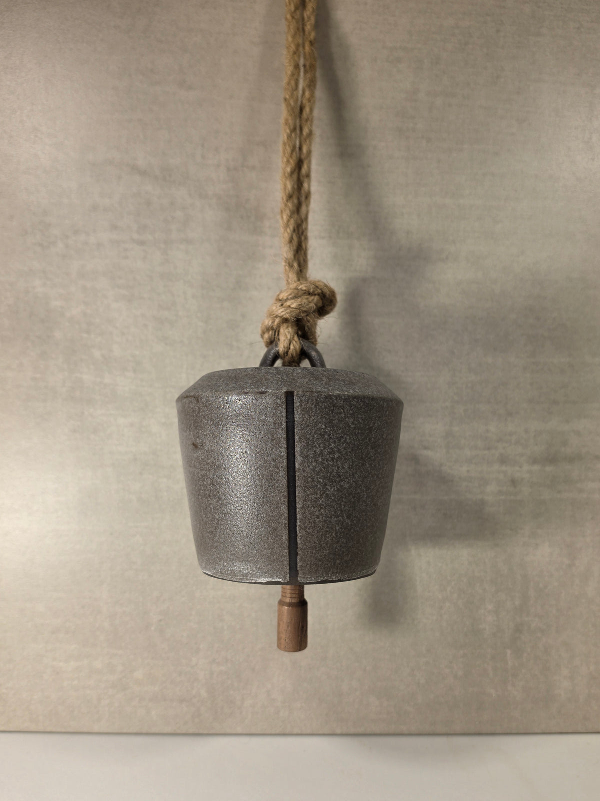 A rustic metal bell with a dark textured finish, suspended by a thick natural jute rope with a wooden weight at the bottom, set against a neutral textured gray background.