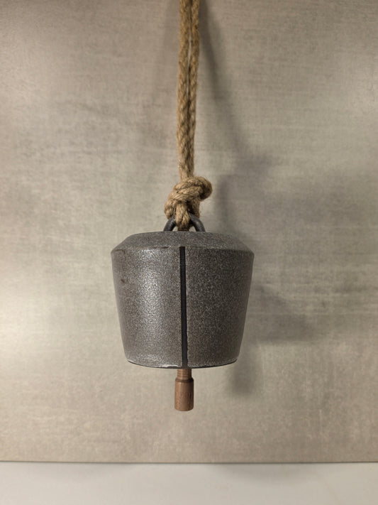 A rustic metal bell with a dark textured finish, suspended by a thick natural jute rope with a wooden weight at the bottom, set against a neutral textured gray background.