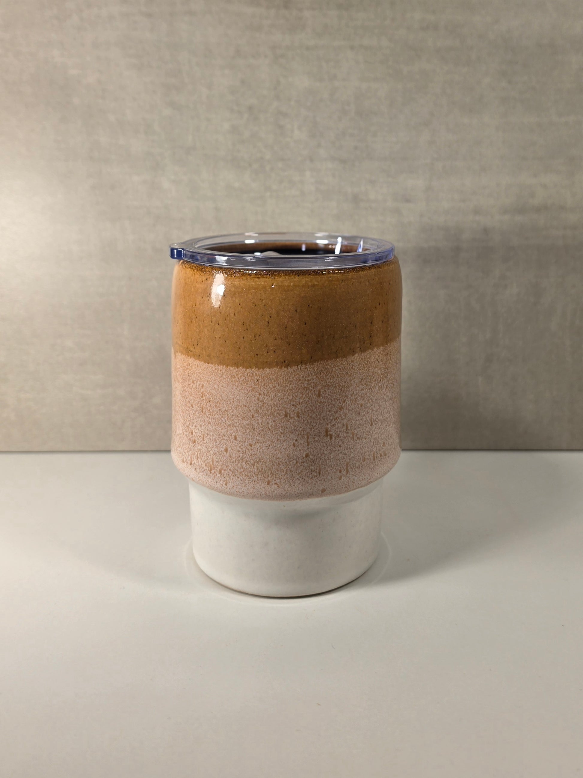 A handmade ceramic tumbler with a three-layered glaze design: a dark brown top band, a speckled light pink middle band, and a matte white bottom base, topped with a clear lid.